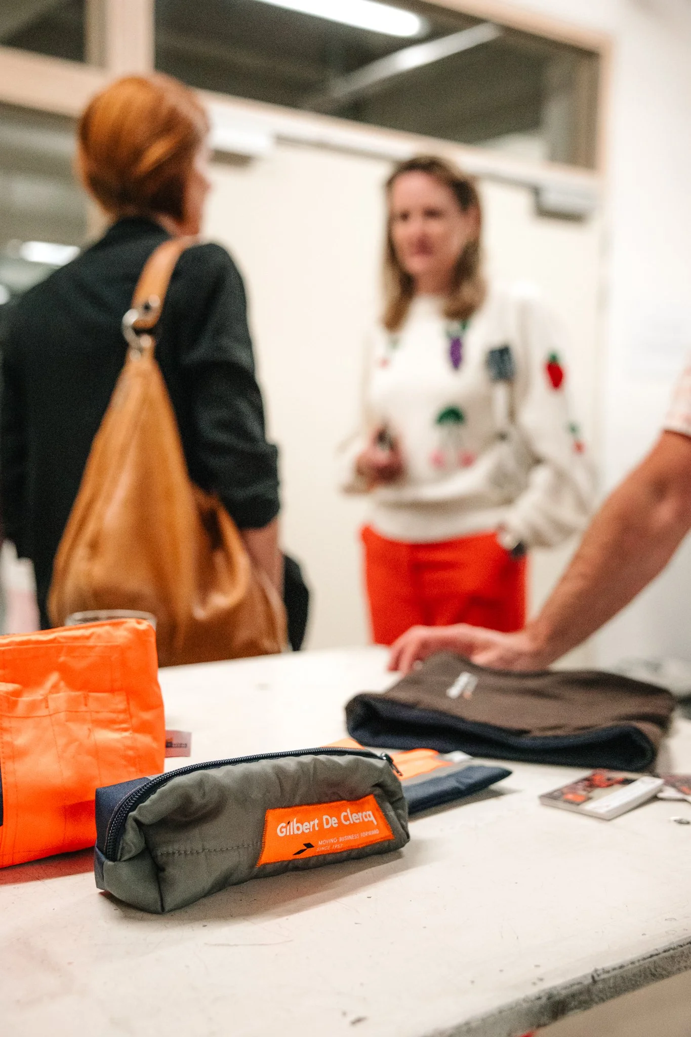 Op de tafel liggen verschillende gepersonaliseerde etuis, waaronder een grijze met oranje textuur, een zwart met oranje label van Gilbert De Clercq, en een feloranje etui. Op de achtergrond praten twee vrouwen, één in een witte blouse met kleurrijke patronen en een oranje rok, en de ander met bruin haar en een zwart jack. De achtergrond is een omgeving die eruitziet als een congres of evenement.