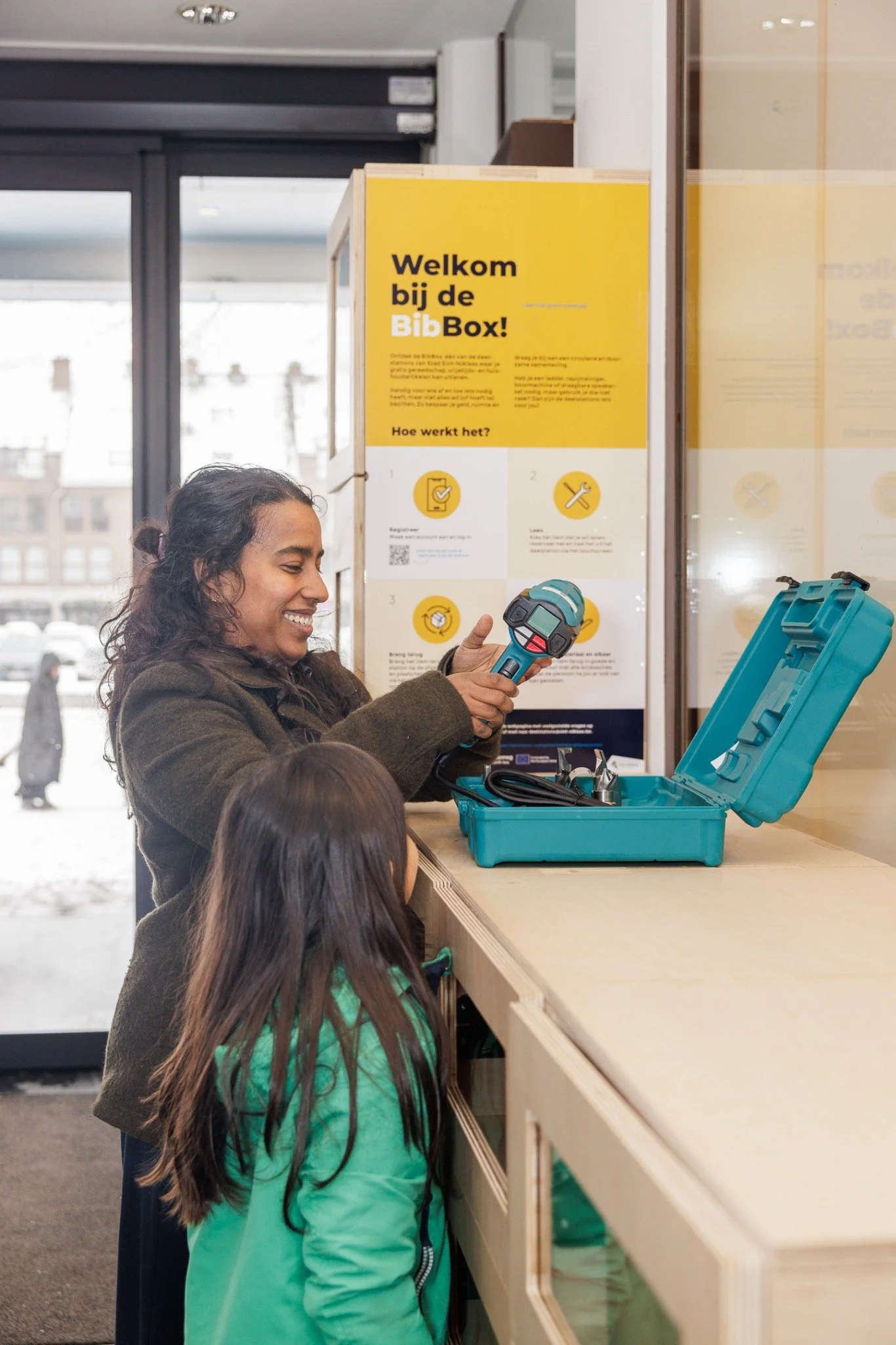 Vrouw en twee kinderen kijken naar een apparaat dat wordt gebruikt voor het meten van temperatuur, bij een informatiestandaard in een openbare ruimte.