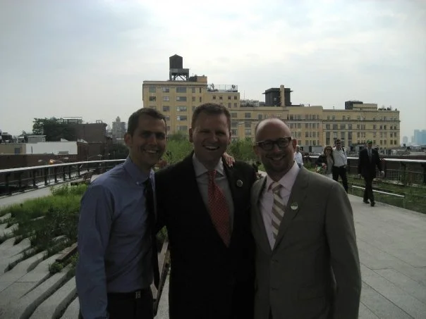 Three men in business attire smiling on a rooftop terrace with city buildings in the background.