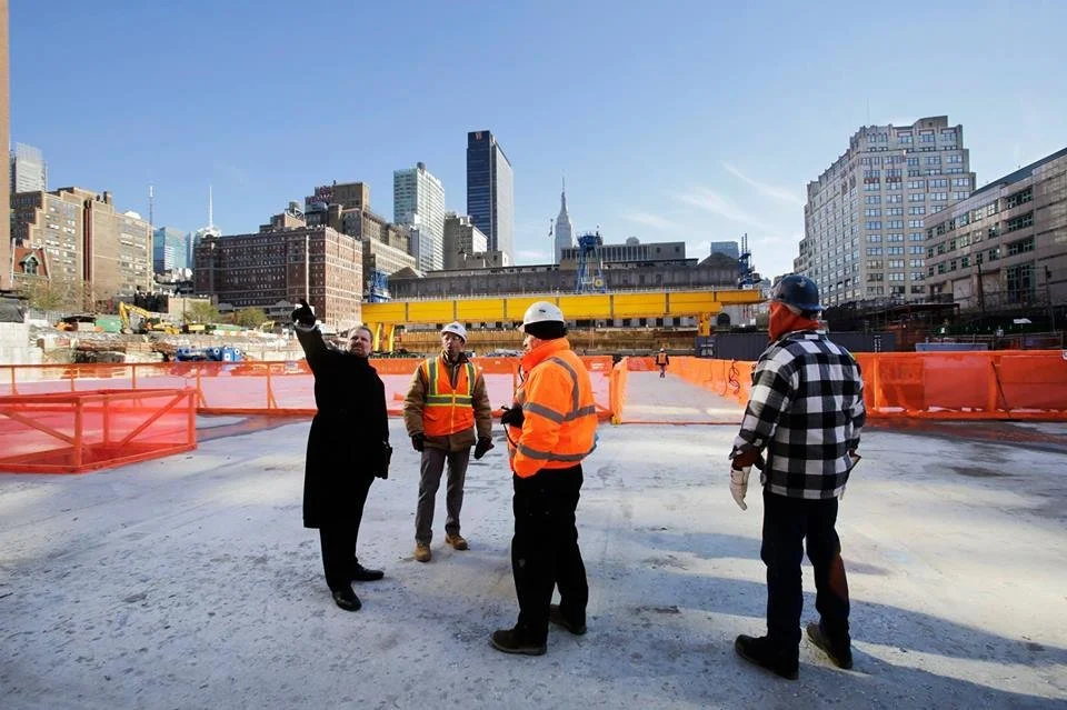 Four construction workers and a man in a suit in a city construction site, with tall buildings and a clear blue sky in the background.