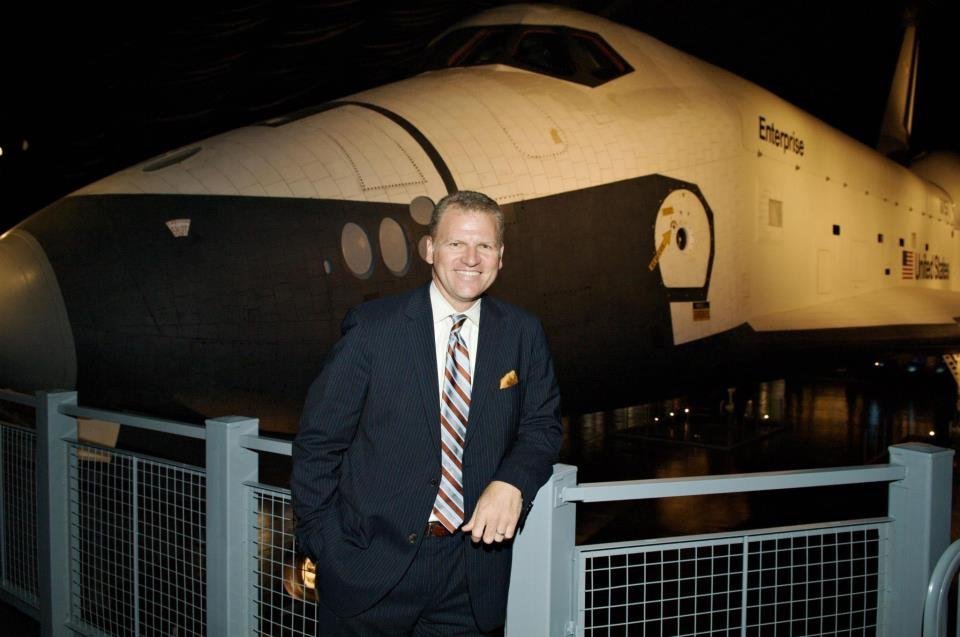 A man in a suit smiling and posing in front of space shuttle Endeavour at a museum exhibit.