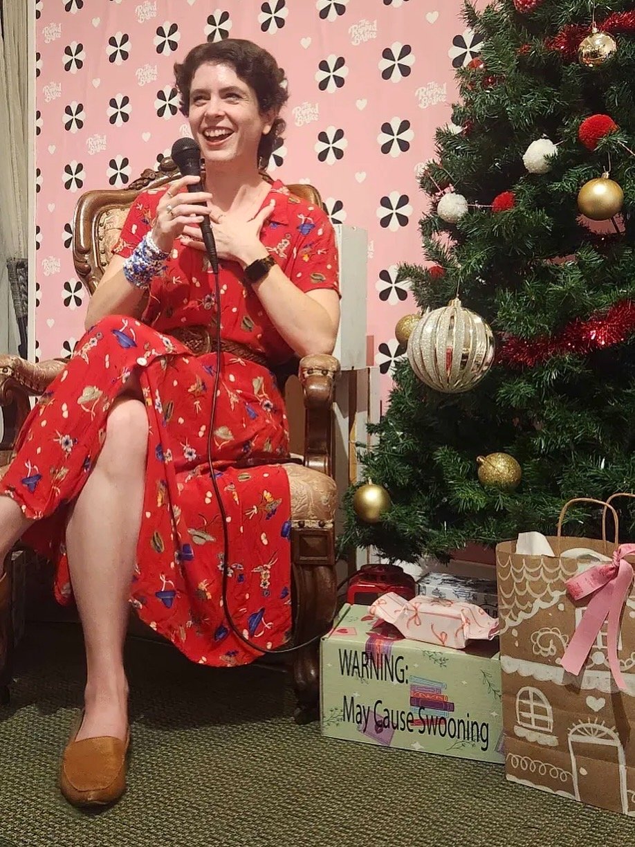 Emily Charlotte, author of Heart Check, smiling and holding a microphone at the Ripped Bodice. Launching her enemies-to-lovers, slowburn, smalltown hockey romance.