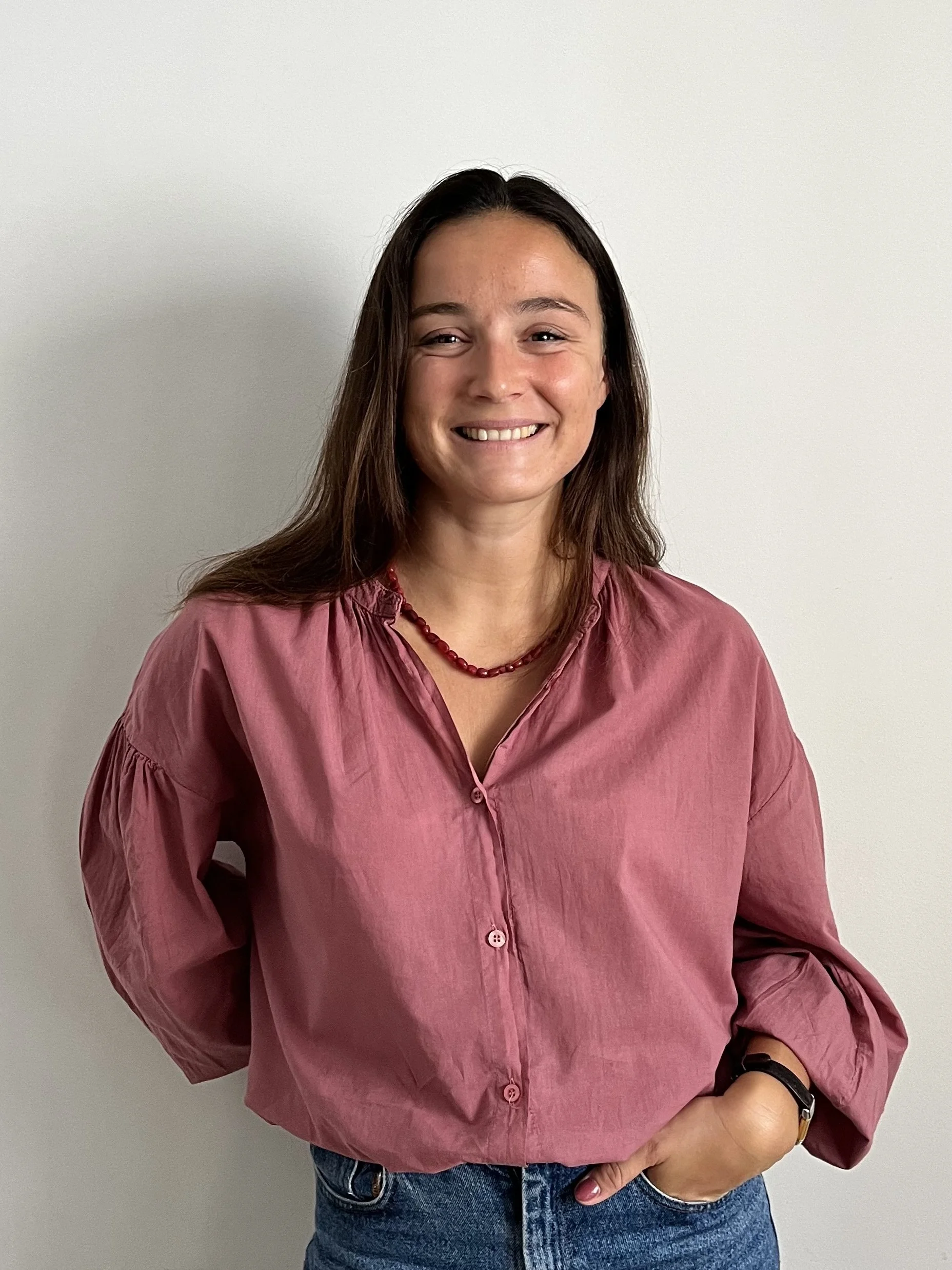 Une femme souriante portant une chemise rose et un collier rouge, posant contre un mur blanc.