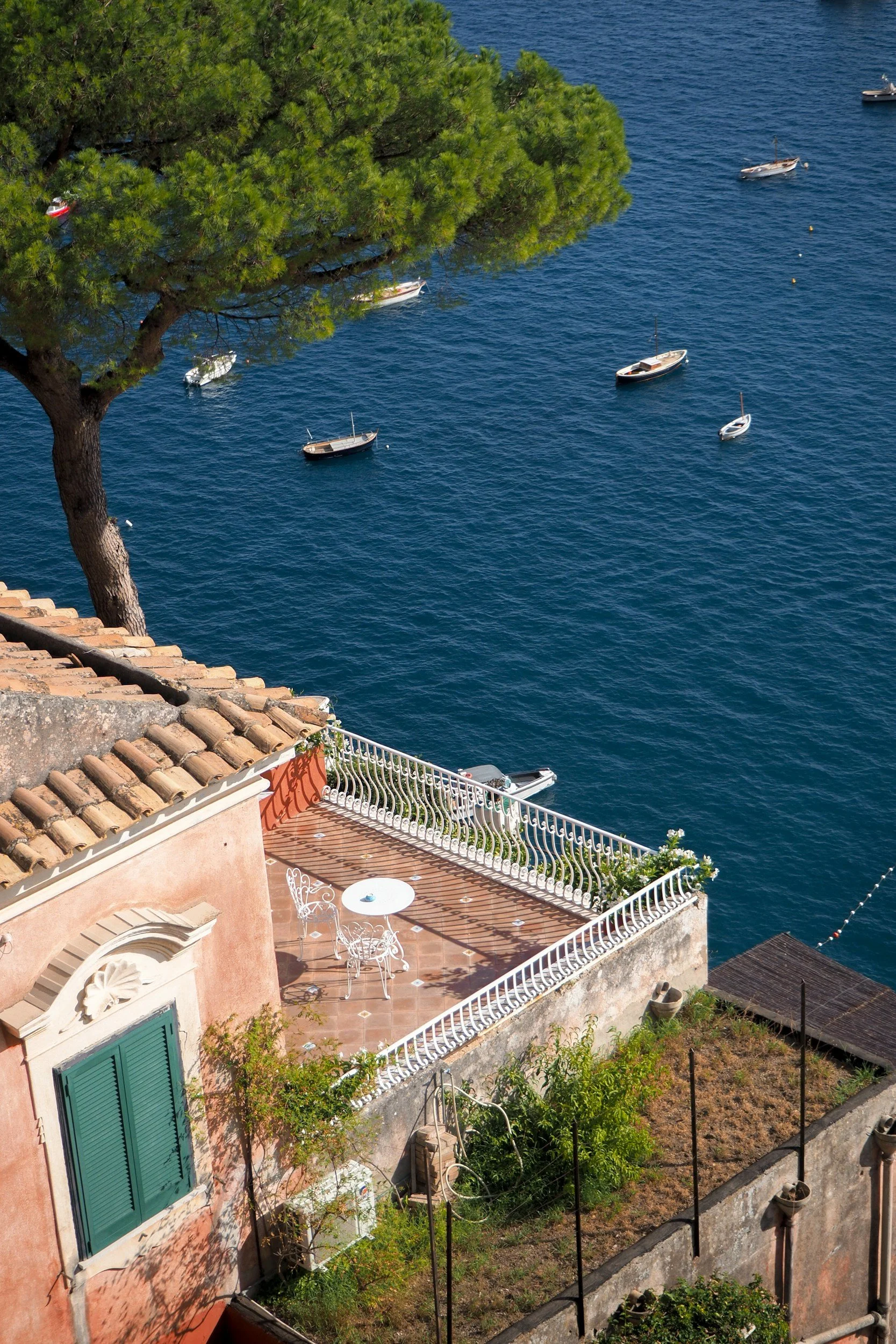 A coastal scene with a pink building featuring a small balcony and outdoor furniture, overlooking a deep blue sea with several anchored sailboats and a large green tree. Amalfi Coast villa.