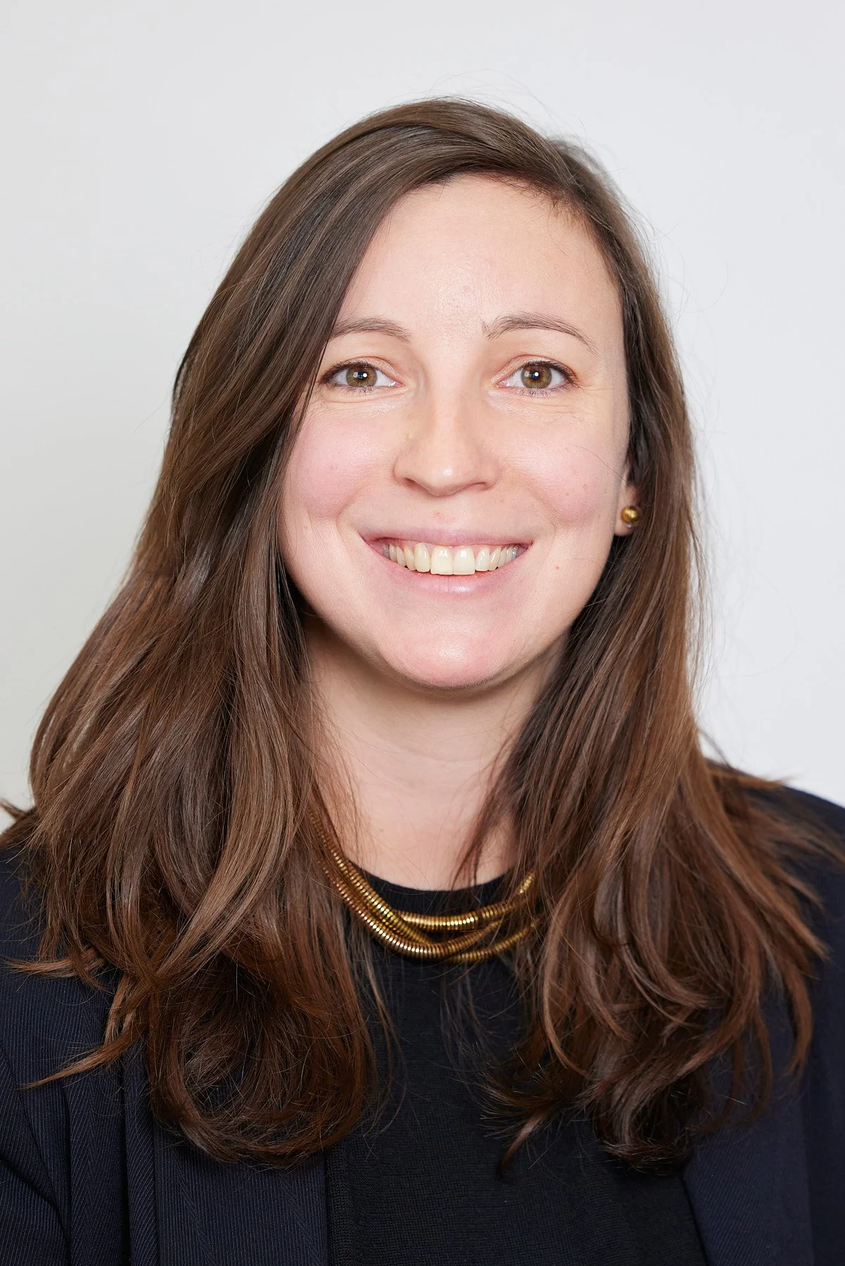 Portrait d'une femme souriante avec des cheveux bruns, portant un collier en or et un vêtement noir, devant un fond blanc.