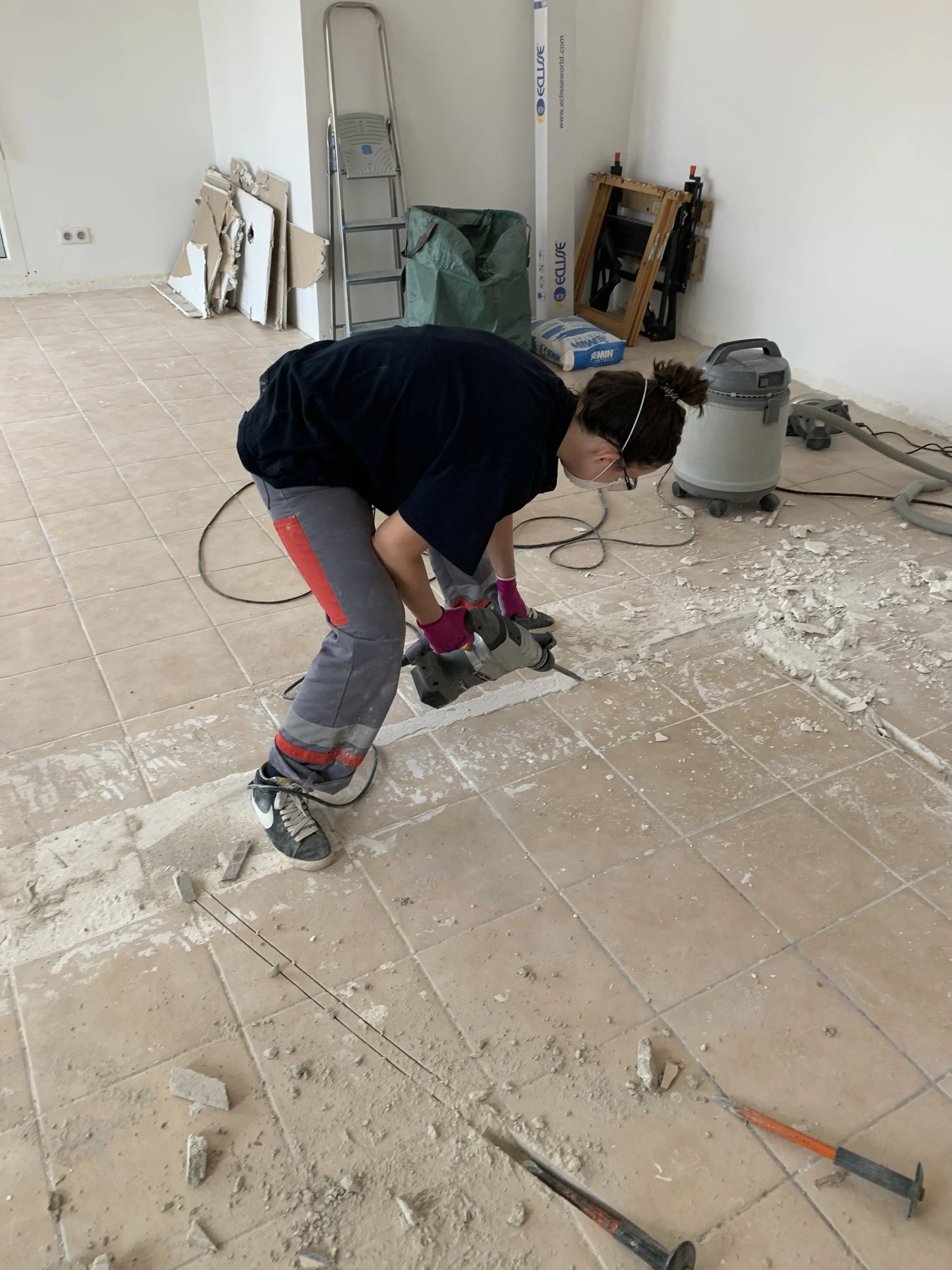 Une personne en train de casser des carreaux de sol avec un marteau-piqueur dans une pièce en rénovation.