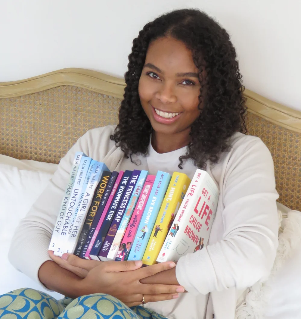 Author Talia Hibbert sitting in bed and and holding all her books in her arms.