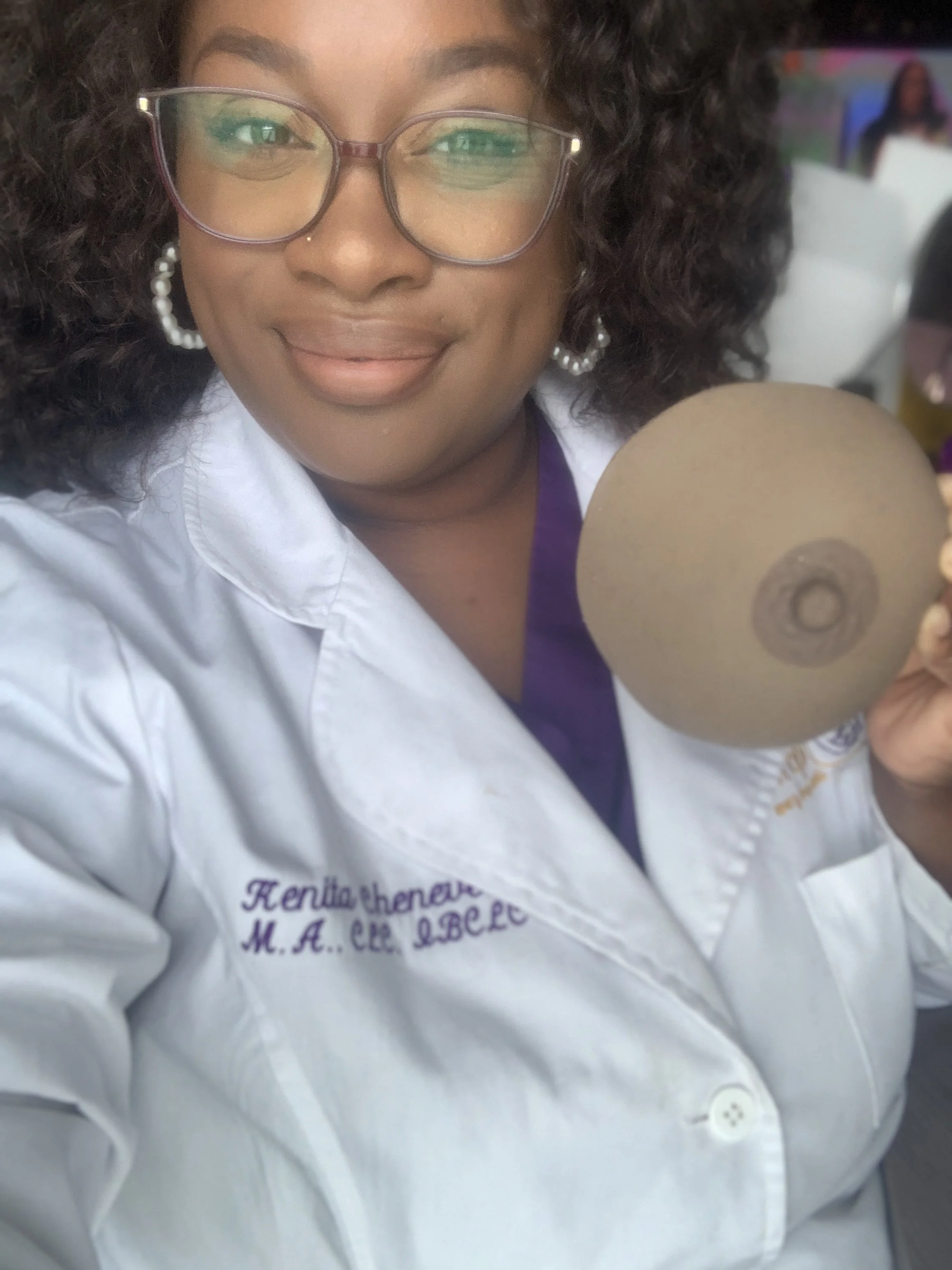 A woman wearing glasses and pearl earrings, dressed in a white coat with embroidered name, smiling and holding a large, round, beige object with a hole in the center.