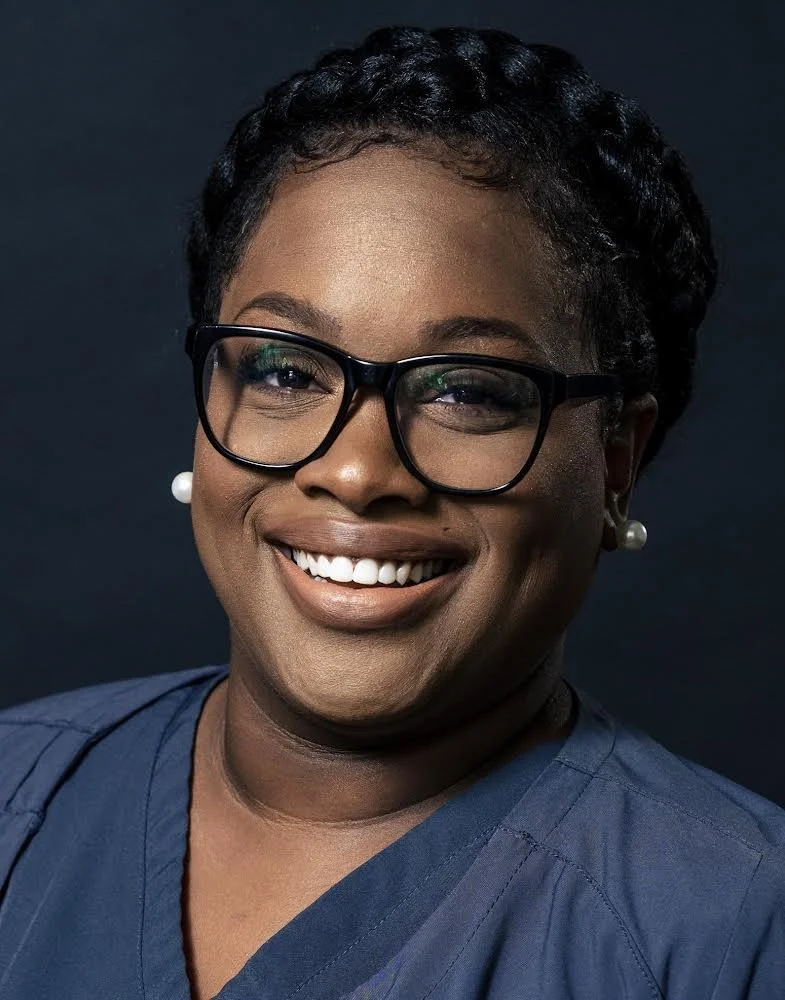 Portrait of a smiling woman with glasses, earrings, and a dark blue top, against a dark background.