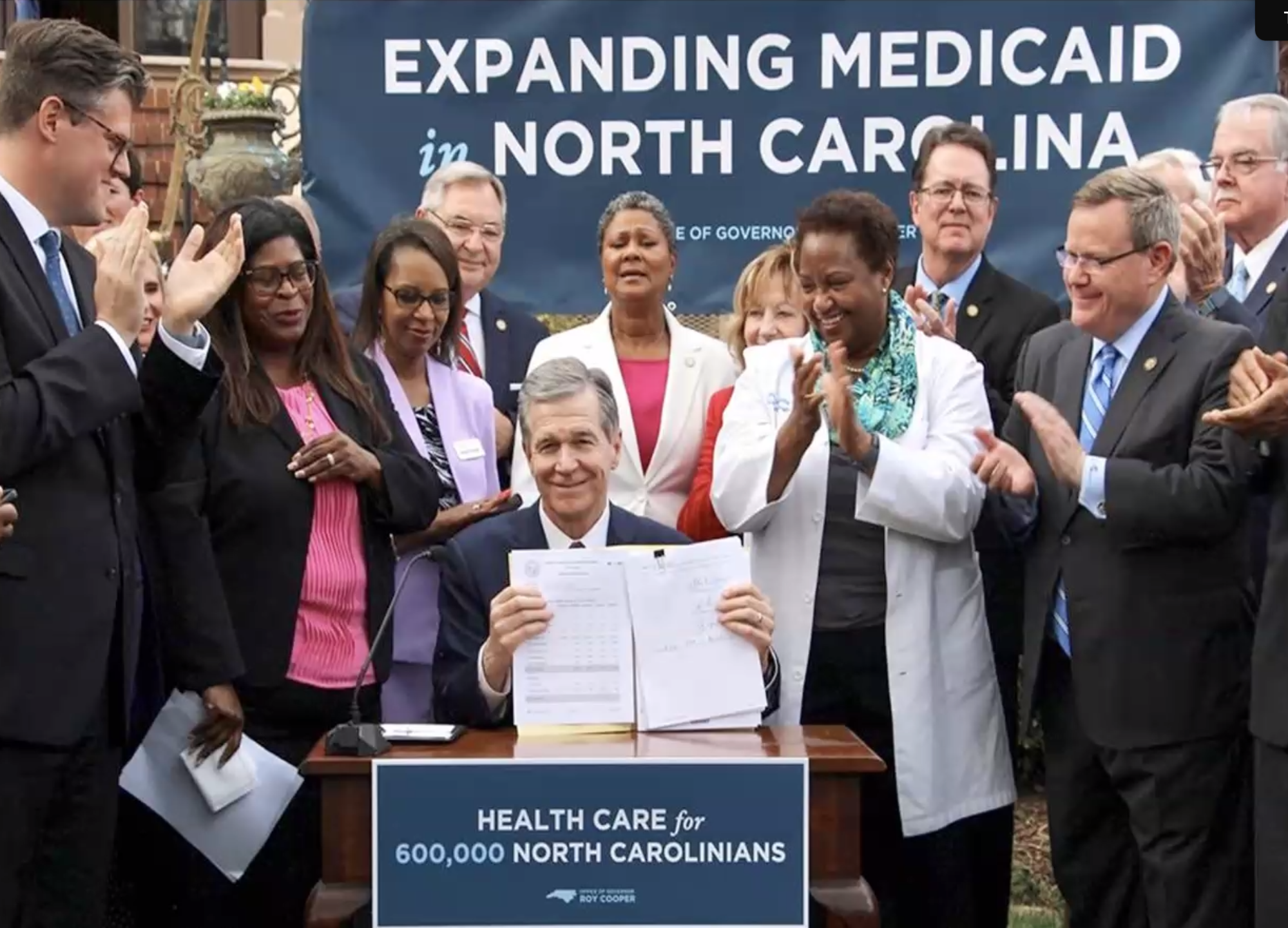 A crowd standing around Roy Cooper smiling and clapping as he holds up legislation. A banner behind him says "Expanding Medicaid in North Carolina" and on the desk in front of him a sign says "Health Care for 600,000 North Carolinians". In the crowd 