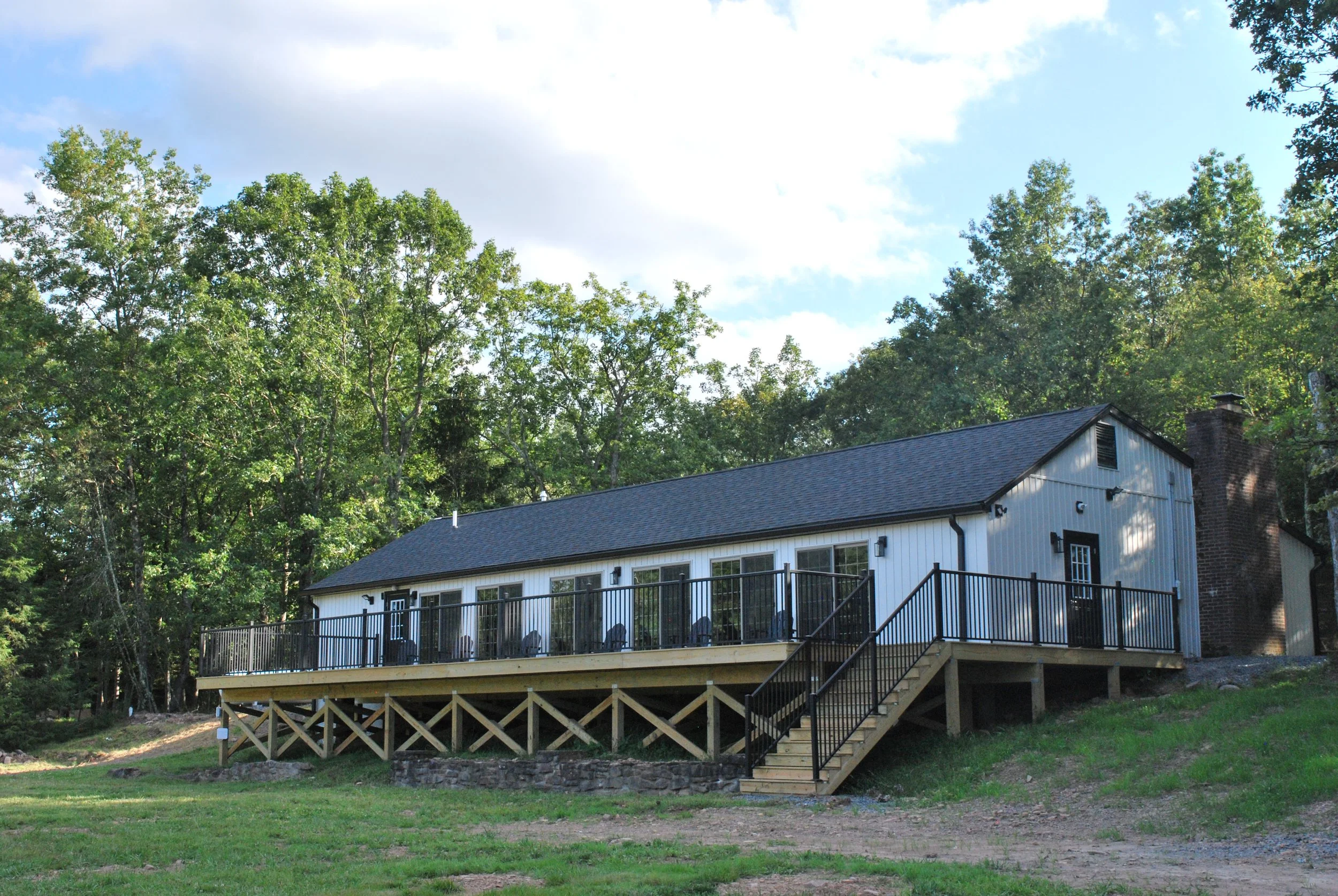 A white house with a black roof and a large porch with black railing, elevated on wooden stilts, with stairs leading up to the porch, situated in a green area with trees in the background and a blue sky with partial clouds.