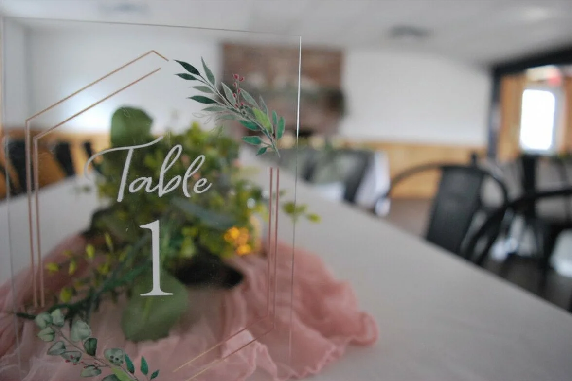 A clear acrylic table number sign with the number 1 and decorated with green leaves and pink accents, placed on a table with a pink tablecloth in a dining room or event space. Wedding venue options.