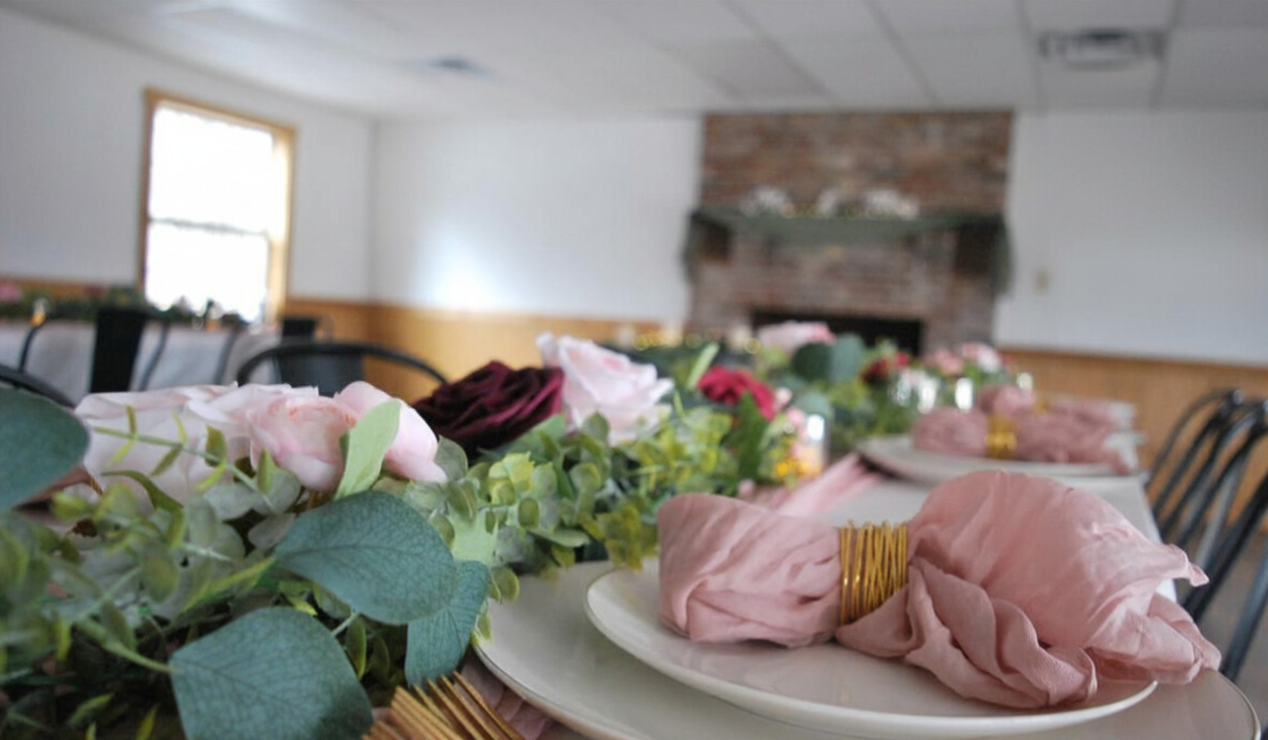 Decorated dining table with pink napkins, floral centerpiece, and place settings in a rustic room with brick fireplace. Wedding venue deor.