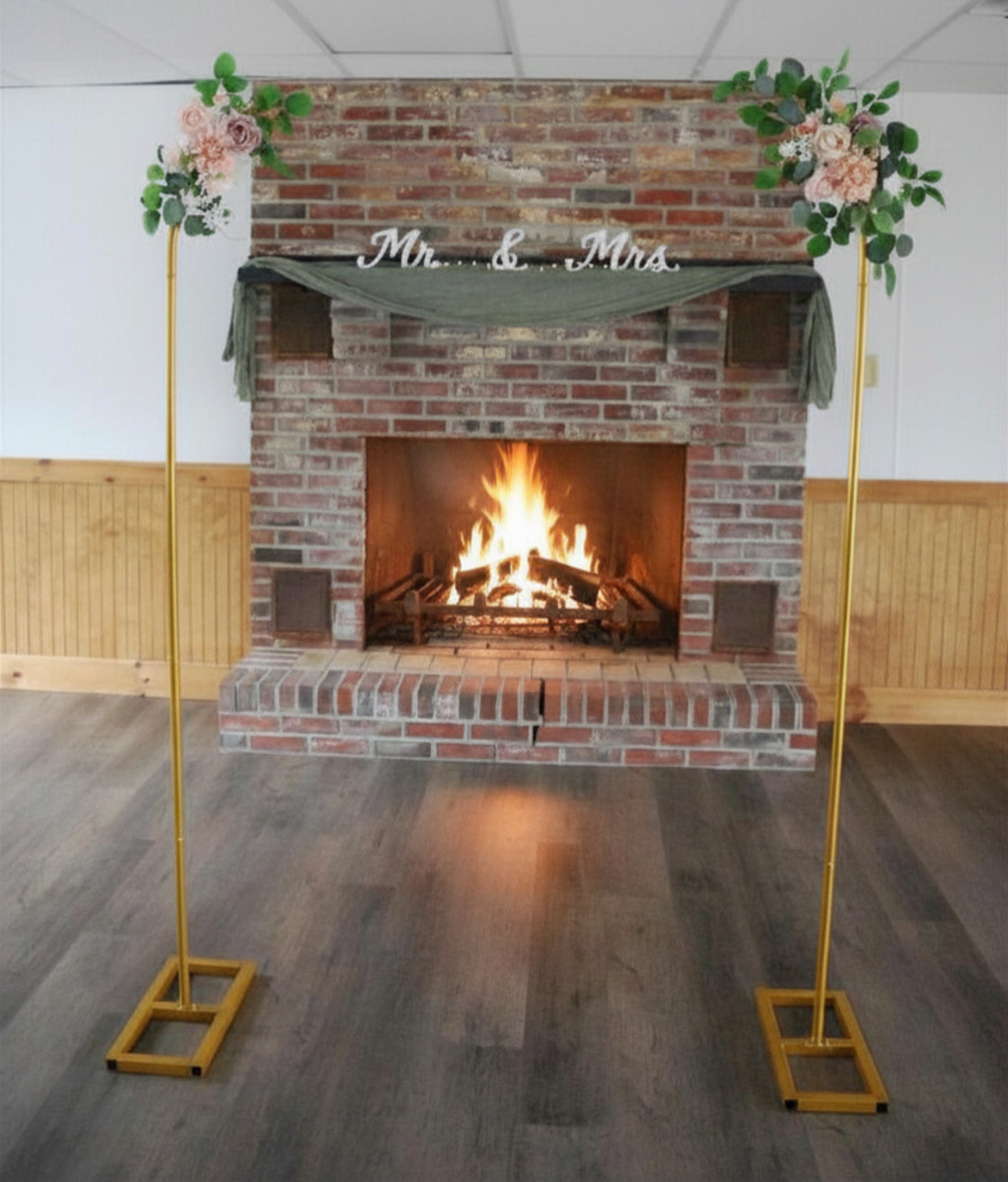 Decoration for a wedding or celebration featuring a brick fireplace with fire, gold stands with floral arrangements on top, and a sign reading 'Mr. & Mrs.' hanging above the fireplace.