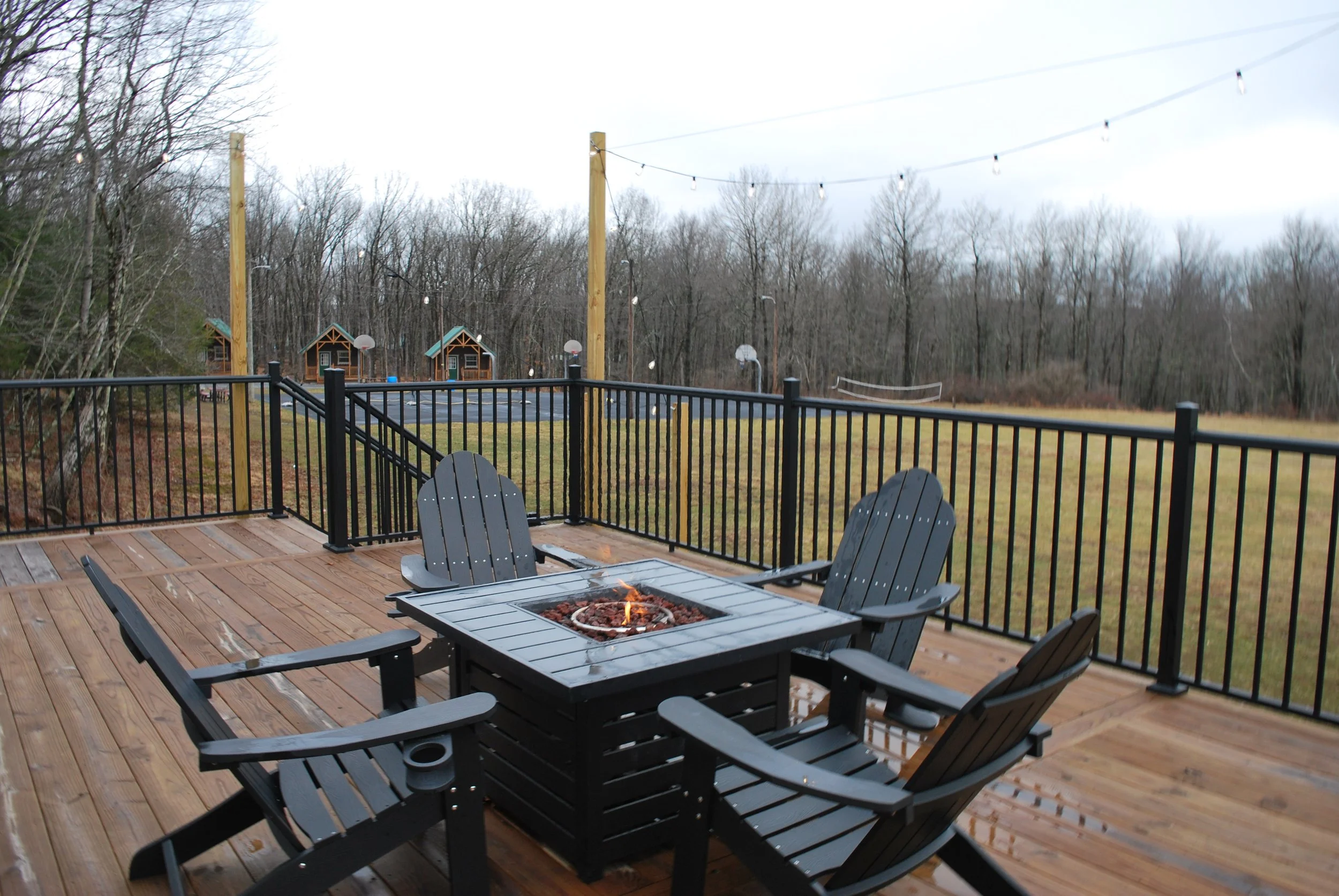 Outdoor wooden deck with a fire pit table surrounded by four black chairs, enclosed by black metal railing, overlooking a grassy field with small structures, basketball hoops, and leafless trees under cloudy sky.