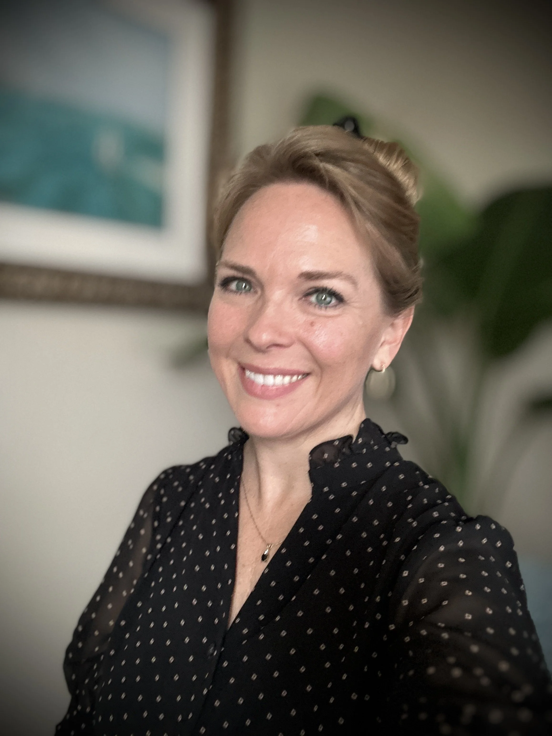 A woman with fair skin, light-colored hair styled back, and blue eyes, smiling for a selfie in a room with a blurred background of a framed picture and a plant.