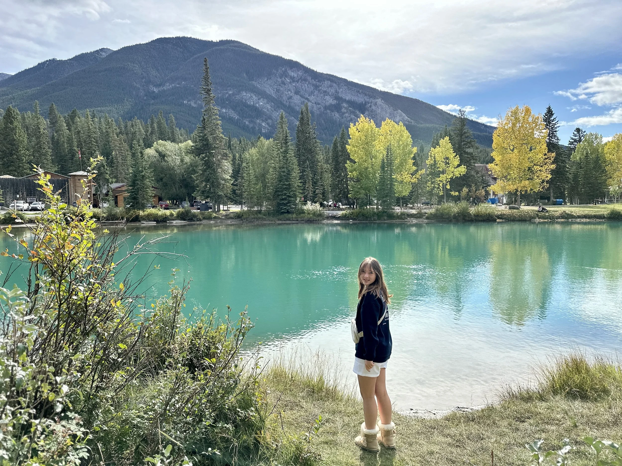 A girl standing at the edge of a lake with turquoise water, surrounded by trees with green and yellow leaves, mountains in the background, and a partly cloudy sky.