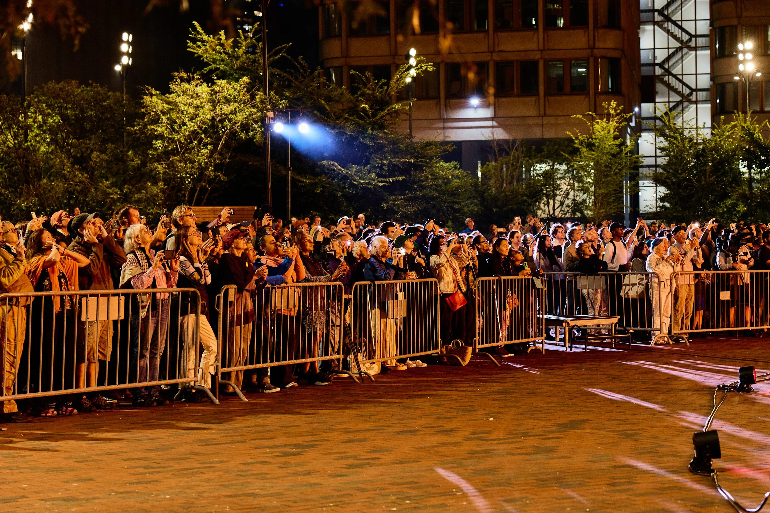 Nighttime outdoor concert with a large audience standing behind a metal barrier, some taking photos or videos with smartphones, illuminated by stage lights, trees, and a modern building in the background.