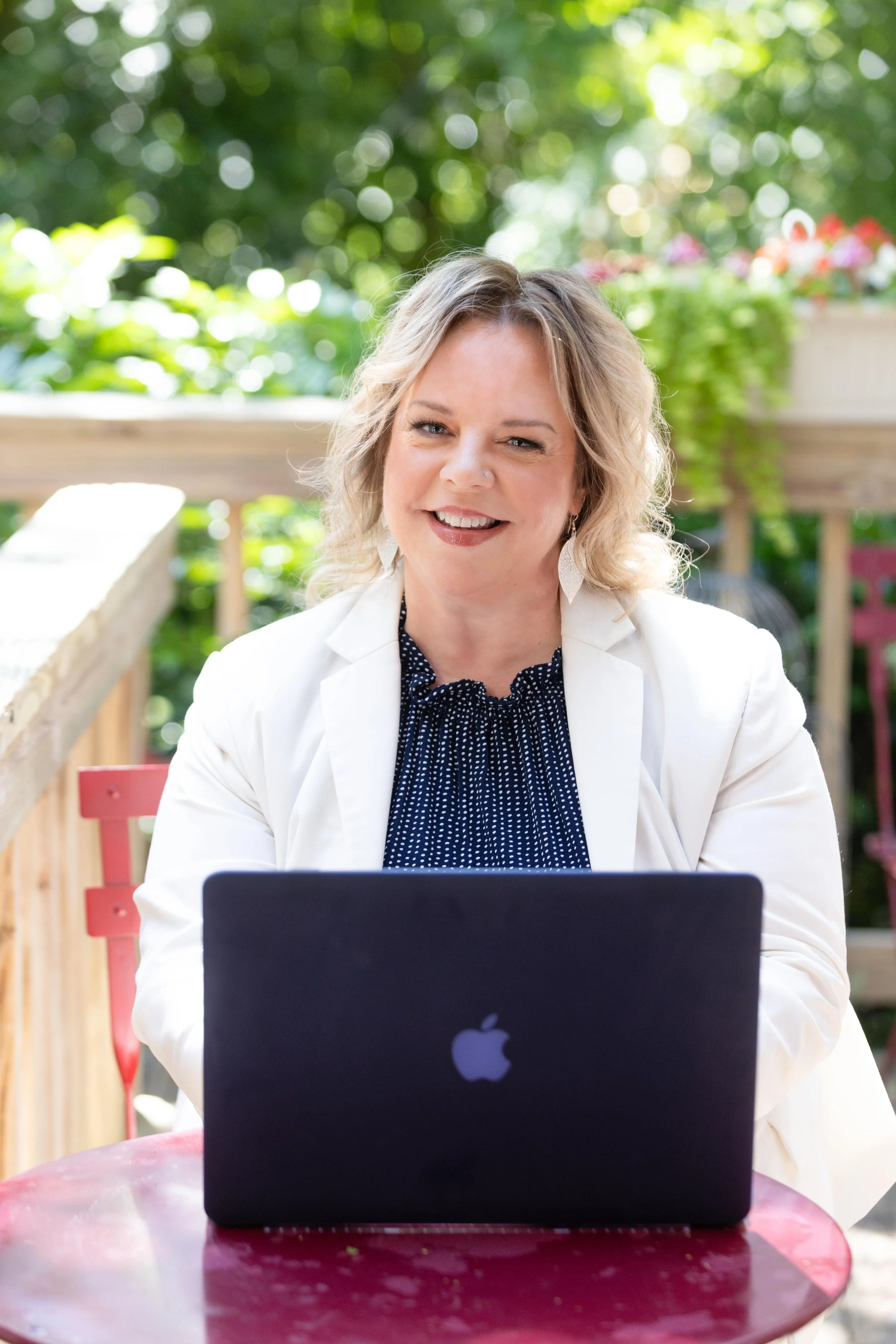 A woman in a white blazer and blue blouse sits outdoors at a red table, smiling while working on a laptop. The background features greenery and flowers, creating a relaxed, professional setting.