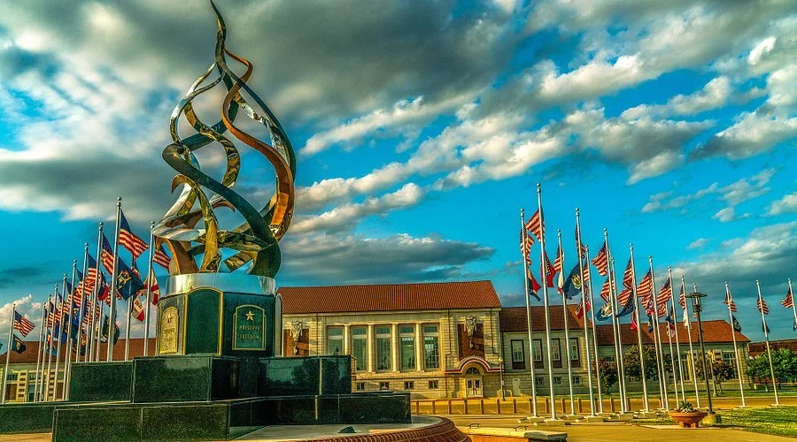 A large, modern sculpture with twisting metallic spirals in front of a building, surrounded by numerous American flags under a partly cloudy sky.