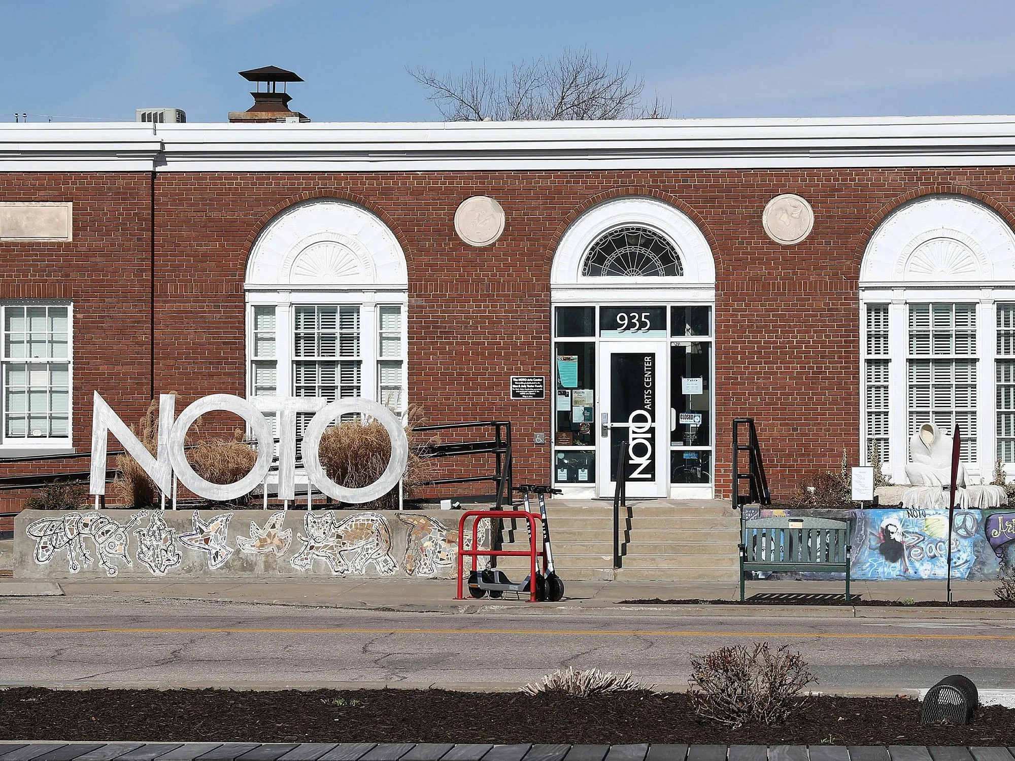 Brick building with white trim around windows, entrance door with number 935, and a sign that reads 'NOTO' in front. There is outdoor art and graffiti on the lower wall, with steps leading up to the entrance.