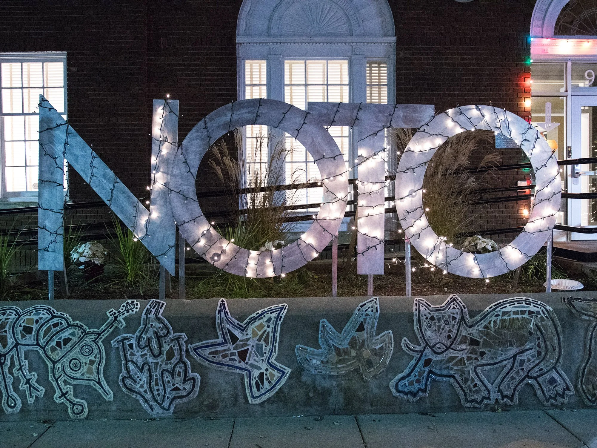 Large illuminated menorah display with decorative drawings of a robot, a rocket, a star, a fox, a dog, and a snake beneath, in front of a brick building with lit windows and holiday decorations.