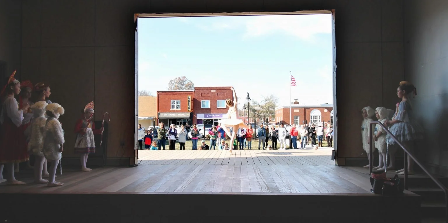 Inside a theater, children in costumes watch a ballet dancer perform on stage with the audience outside in a small town