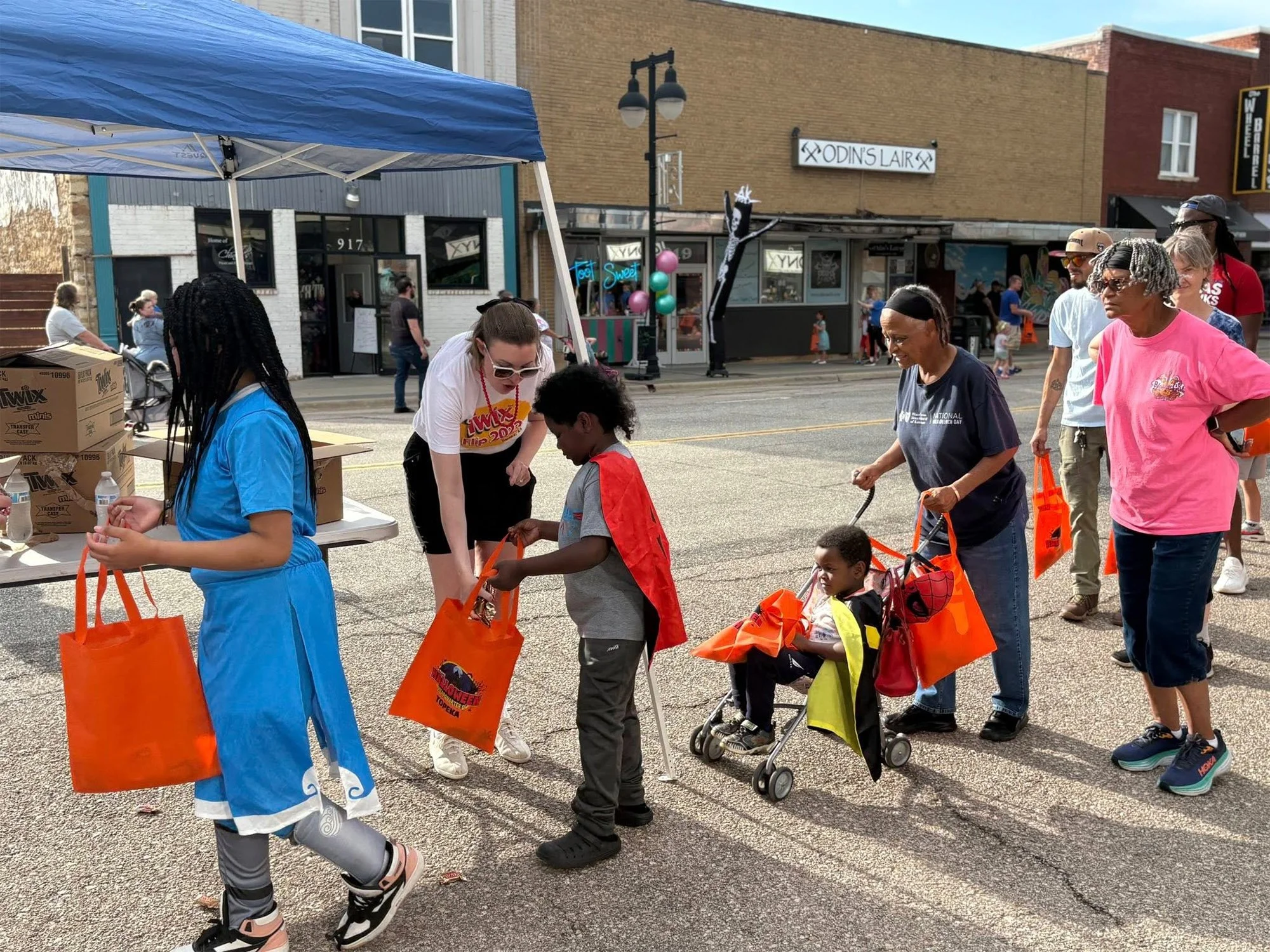People participating in an outdoor event, with some handing out supplies to children, some in wheelchairs, and several wearing colorful clothing, on a city street with shops and banners in the background.