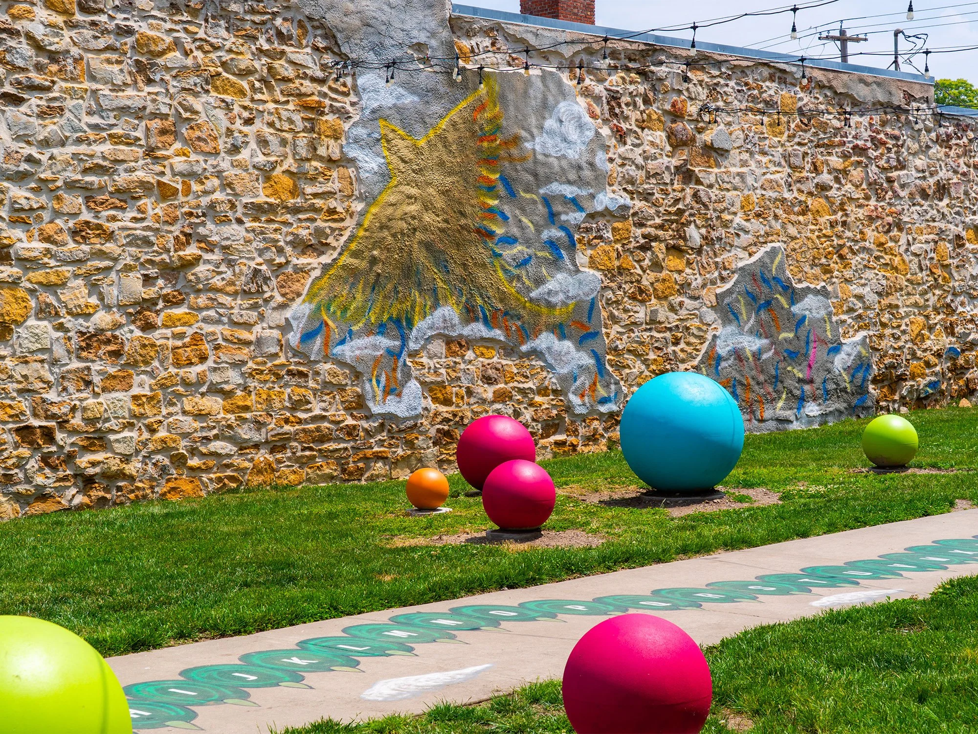 Colorful outdoor art installation with large painted balls in pink, green, orange, and blue on a grassy area next to a stone wall featuring mural artwork of a mountain with clouds and rainbow accents.