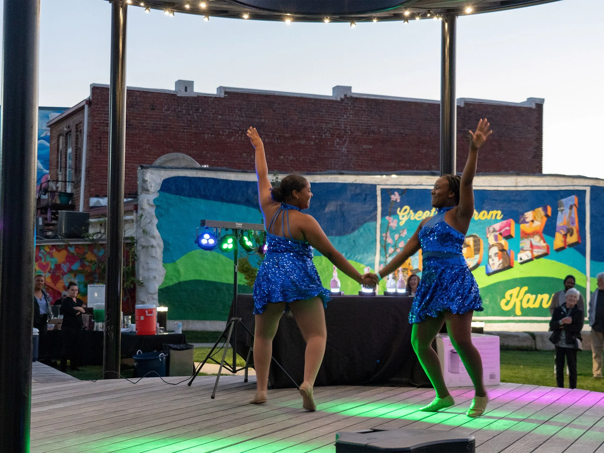 Two women in blue sequined dresses dancing on a wooden stage outdoors during an event, with a colorful mural in the background. Several people are watching, and there are green and purple stage lights.
