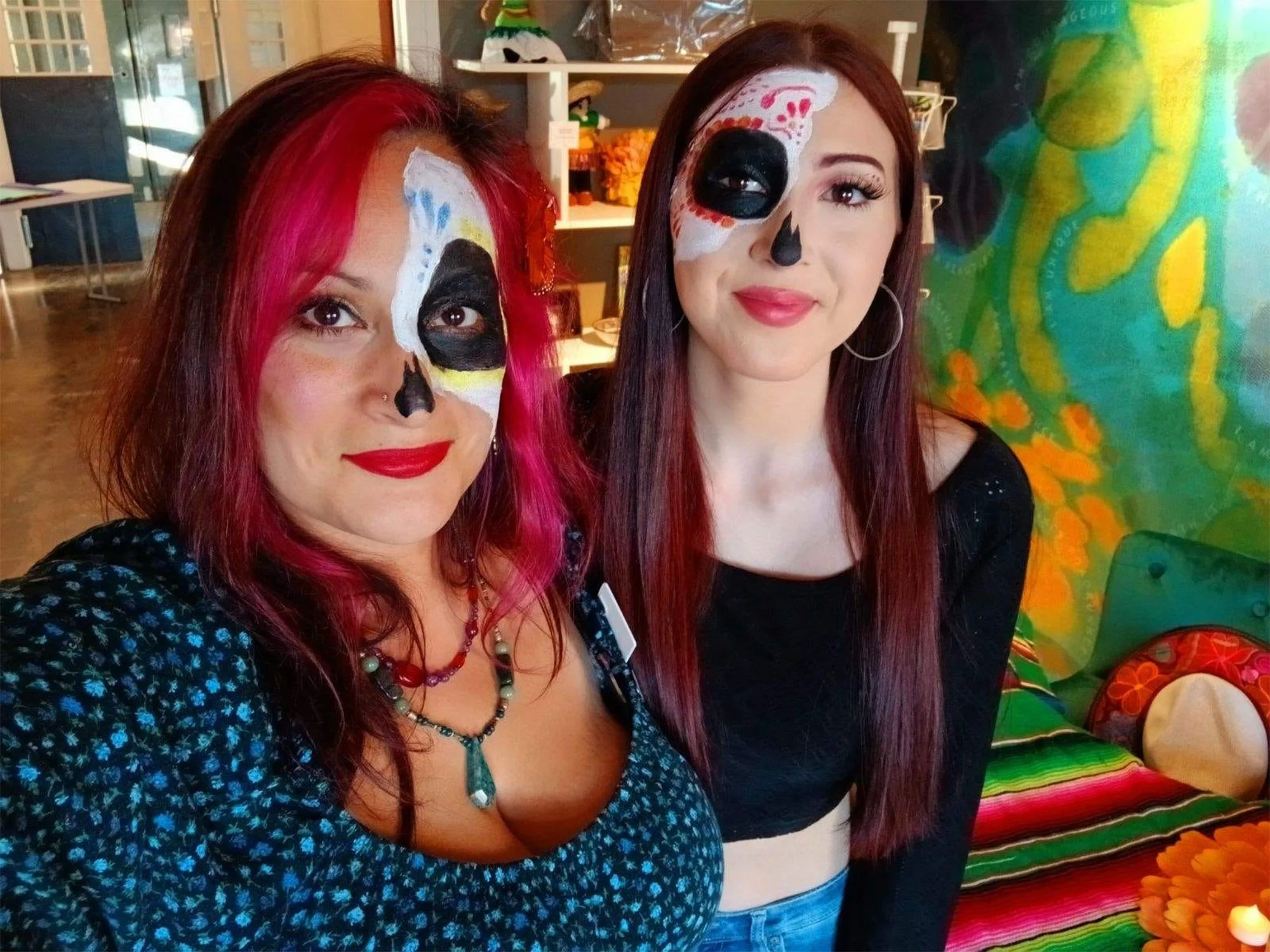 Two women with face painted as sugar skulls, standing inside a colorful room decorated with vibrant Mexican-themed decorations for Día de los Muertos.