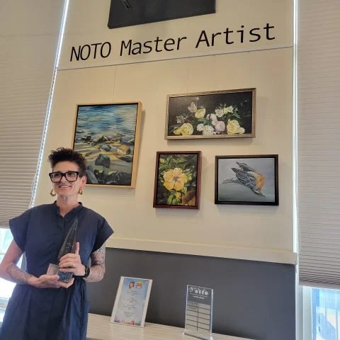 Young woman with glasses and tattoos holding an award trophy at the NOTO Master Artist exhibition, with four paintings on the wall behind her.