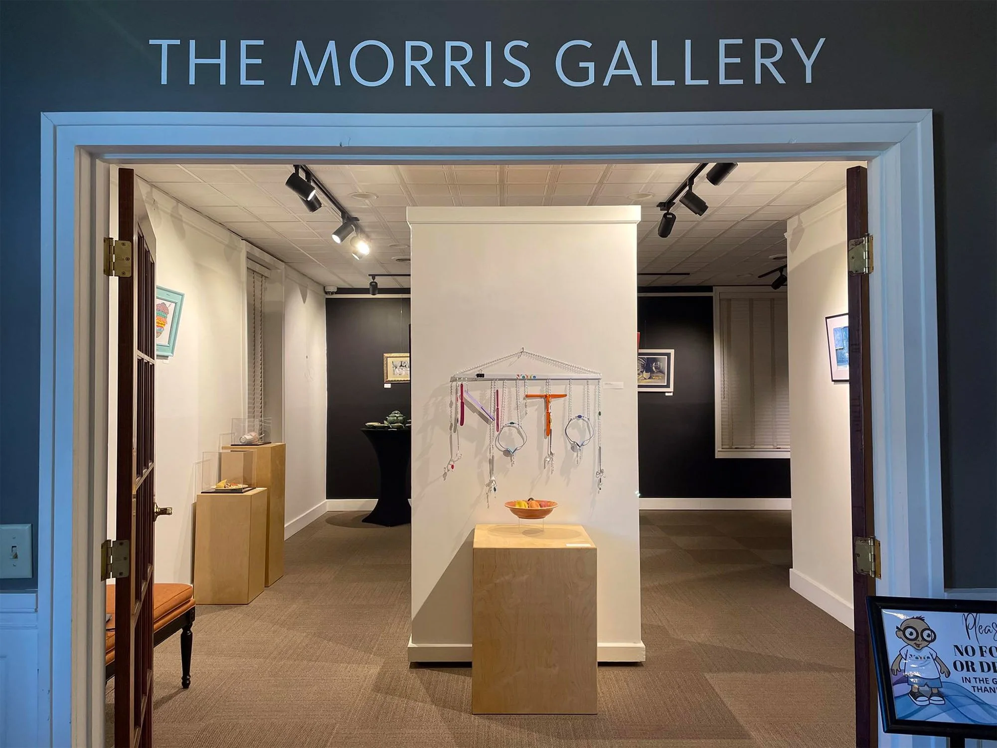 Entrance to The Morris Gallery with displayed artwork, a wooden bench, and a sign about gallery policies.