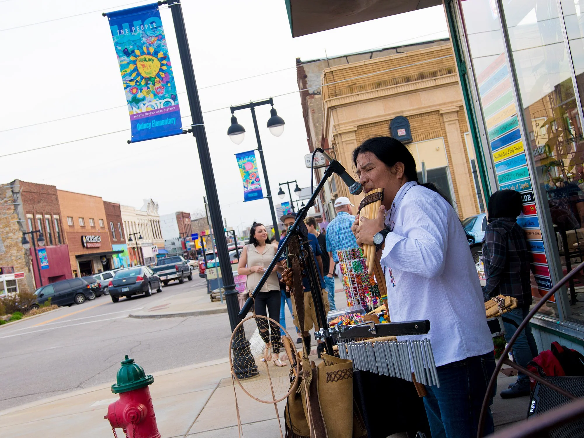 A street performer playing a pan flute and singing into a microphone on a busy city sidewalk, with pedestrians and colorful storefronts in the background.