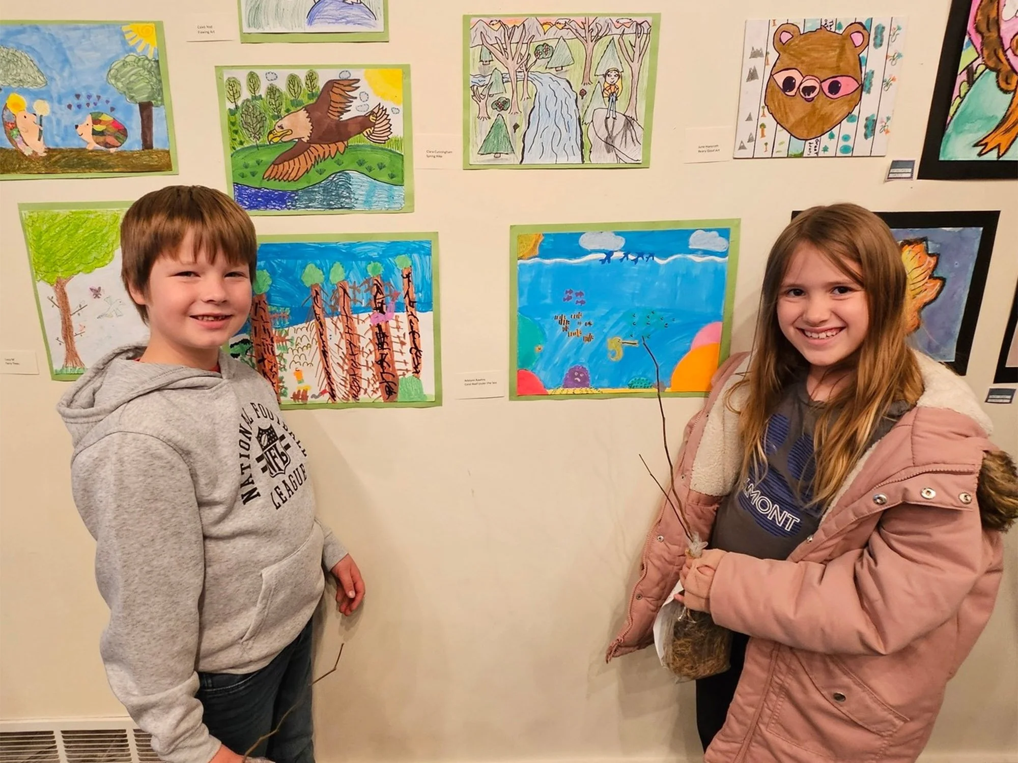 Two children, a boy and girl, standing in front of a wall displaying colorful artwork of nature scenes and animals; the boy is wearing a gray hoodie and the girl a pink jacket, both smiling.