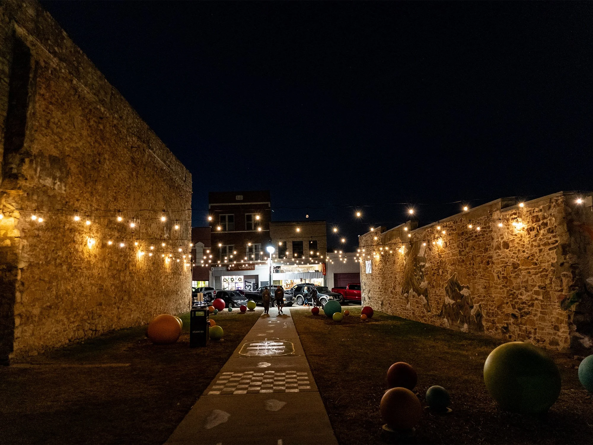 Night view of a small urban alleyway decorated with string lights, with large decorative spheres on the ground and a mural on the right wall, leading toward a street with parked cars and a building in the background.