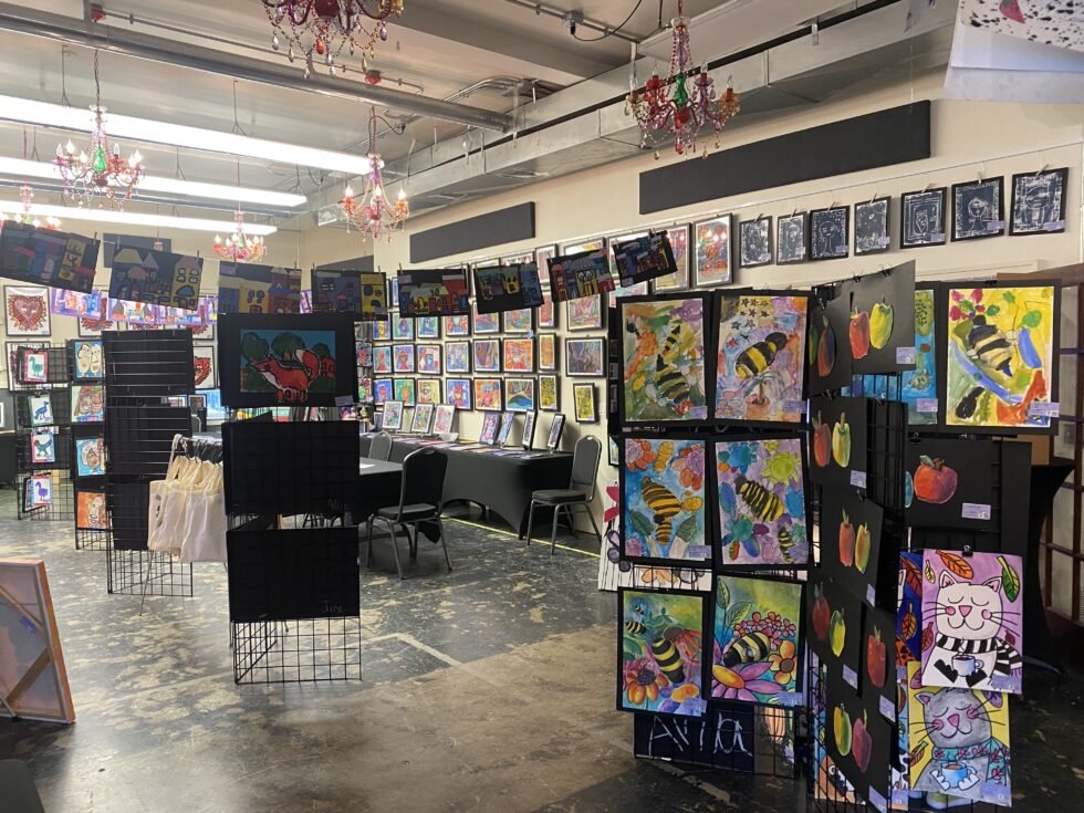 Art gallery with colorful paintings, including bees and fruit, displayed on walls and on panels, with chandeliers hanging from the ceiling.