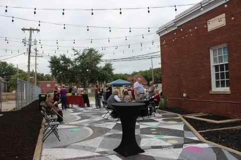 People gathered at an outdoor event on a decorated patio with string lights, tables, and chairs next to a red brick building.