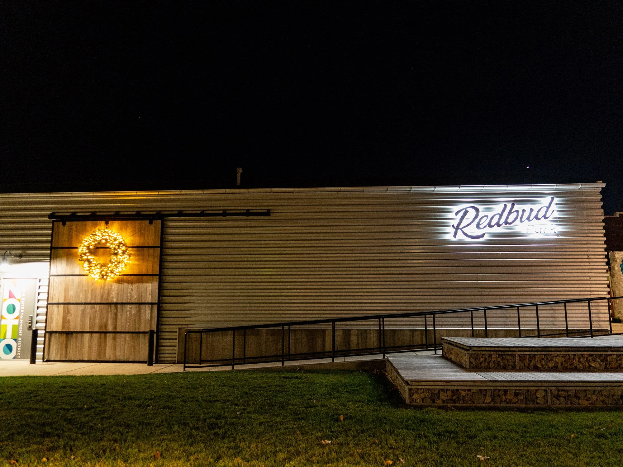 Nighttime exterior view of a building with a sign that says 'Redbud,' a wooden door decorated with a Christmas wreath, and a small ramp leading to the entrance.