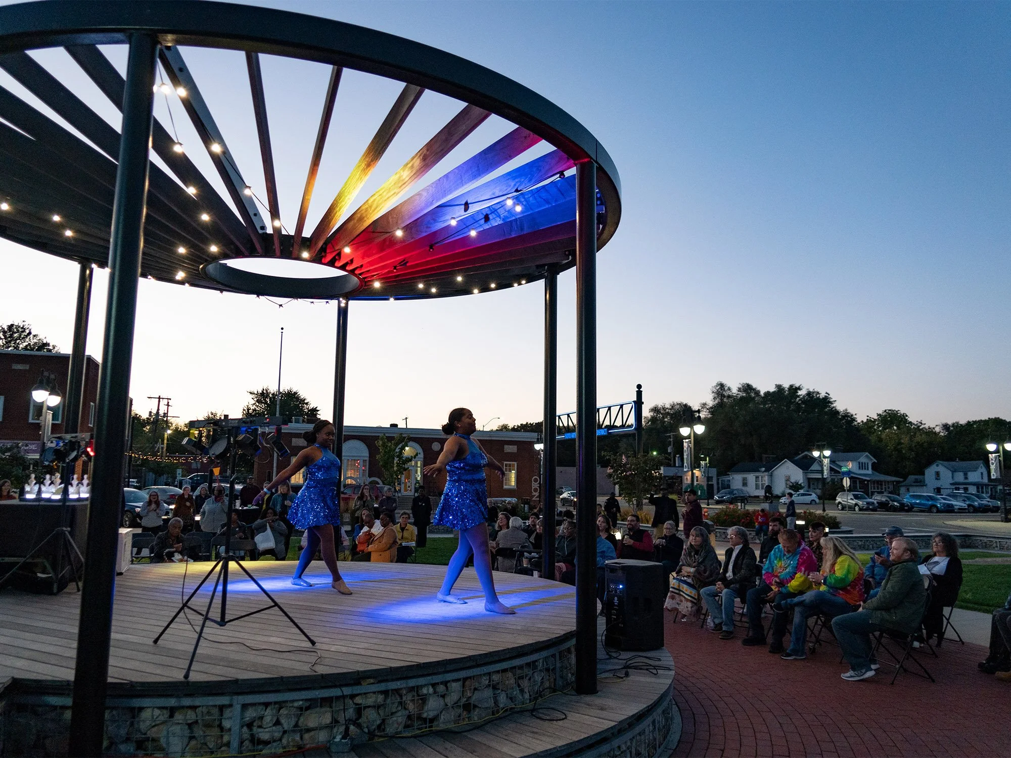 Three performers in blue sequined dresses dancing on a small outdoor stage during a concert at dusk, with an audience seated in front and a cityscape in the background.