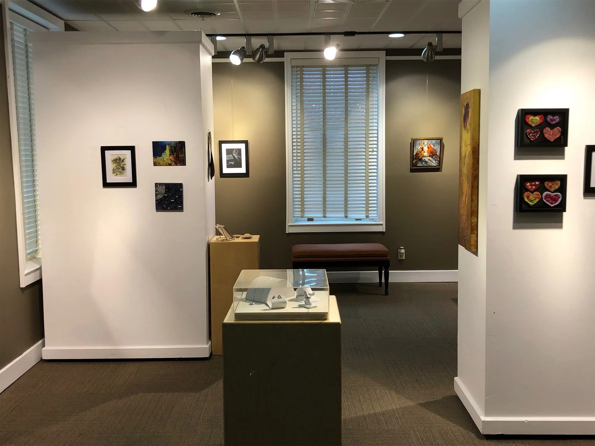 An art gallery with paintings and artwork displayed on white and green walls, a glass display case in the center with jewelry inside, a bench near a window with closed blinds, and track lighting illuminating the art pieces.