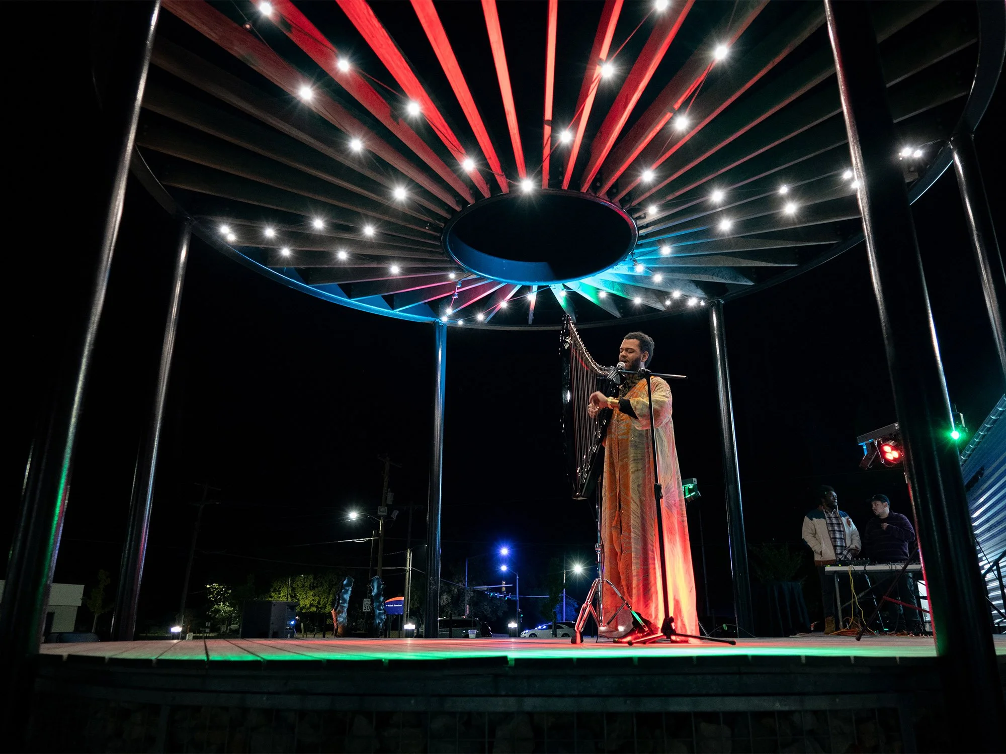A man in traditional attire playing a harp on an outdoor stage at night, with colorful lights on a circular canopy overhead, and two other musicians in the background.