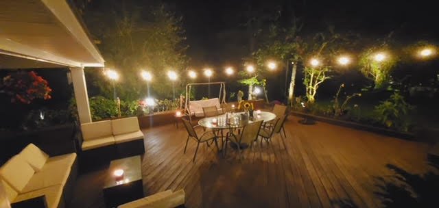 Outdoor patio at night with string lights, a round table with chairs, a wooden swing, and seating area on a wooden deck.