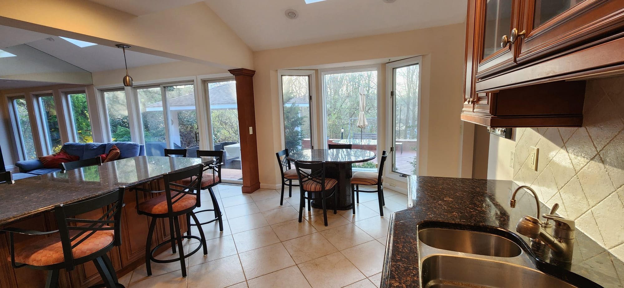 Spacious kitchen with granite countertops, wooden cabinets, dining table, bar stools, and large windows overlooking a patio and greenery.