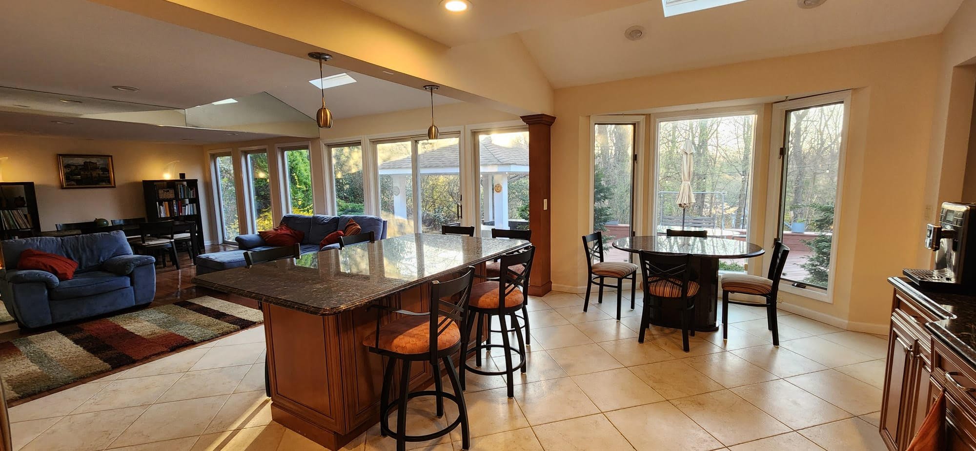 Modern open-concept kitchen and living area with large windows. The kitchen features a granite island with bar stools and adjacent dining area. The living room has a sofa, bookshelves, and a colorful rug. Views of a garden are visible through the win