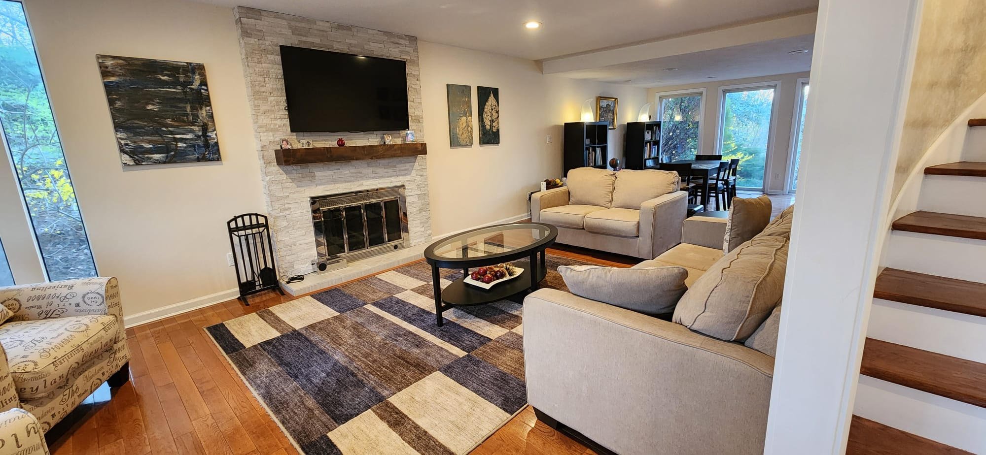 Modern living room with beige sofas, a glass coffee table, a wall-mounted TV above a fireplace, abstract wall art, a plaid area rug, and a dining area with large windows in the background.