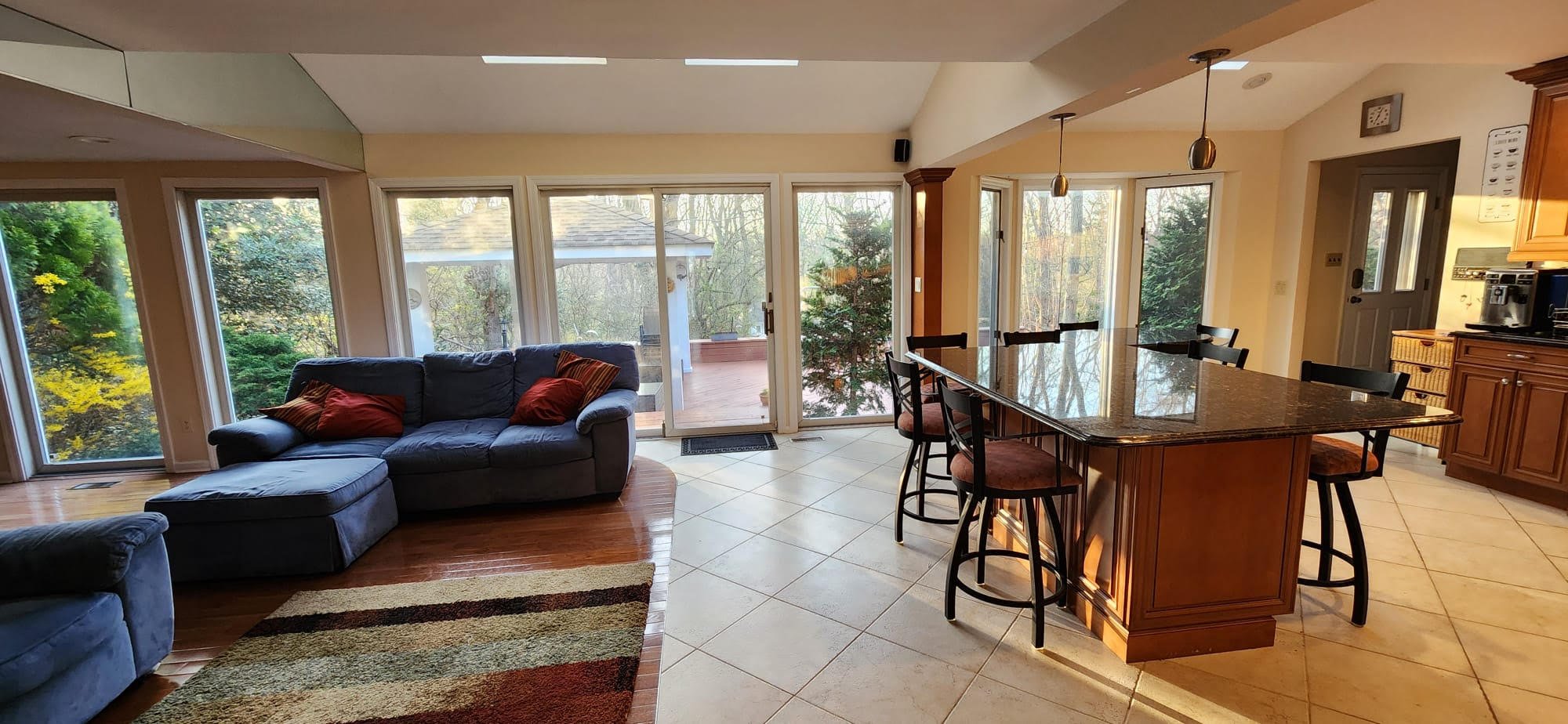 Modern kitchen and living area with large windows, a blue sectional sofa, wooden floors, a striped area rug, and a granite kitchen island with bar stools.