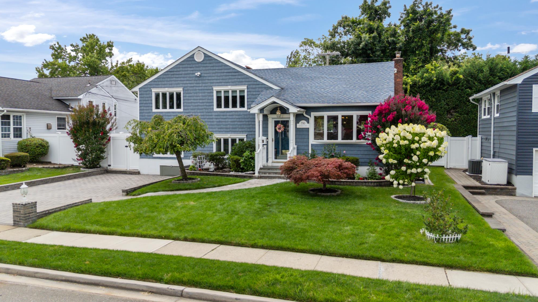 A blue house with a well-maintained front yard featuring colorful bushes and trees, a brick-paved driveway, and a white fence.