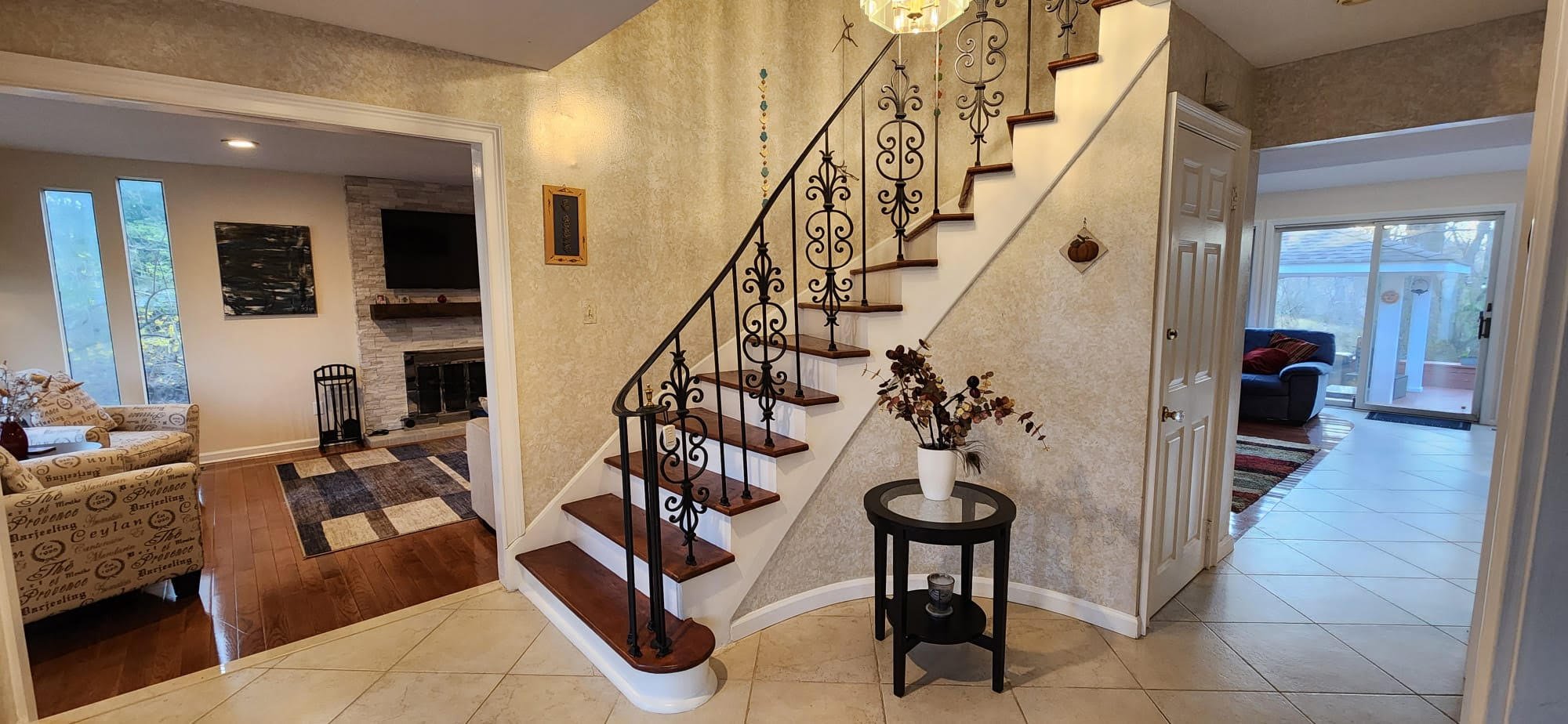 Interior of a home featuring a staircase with ornate iron railings, a living room with a fireplace and television, and decorative plants on a round table. The room includes tiled flooring and a view of a sunroom with sliding glass doors.