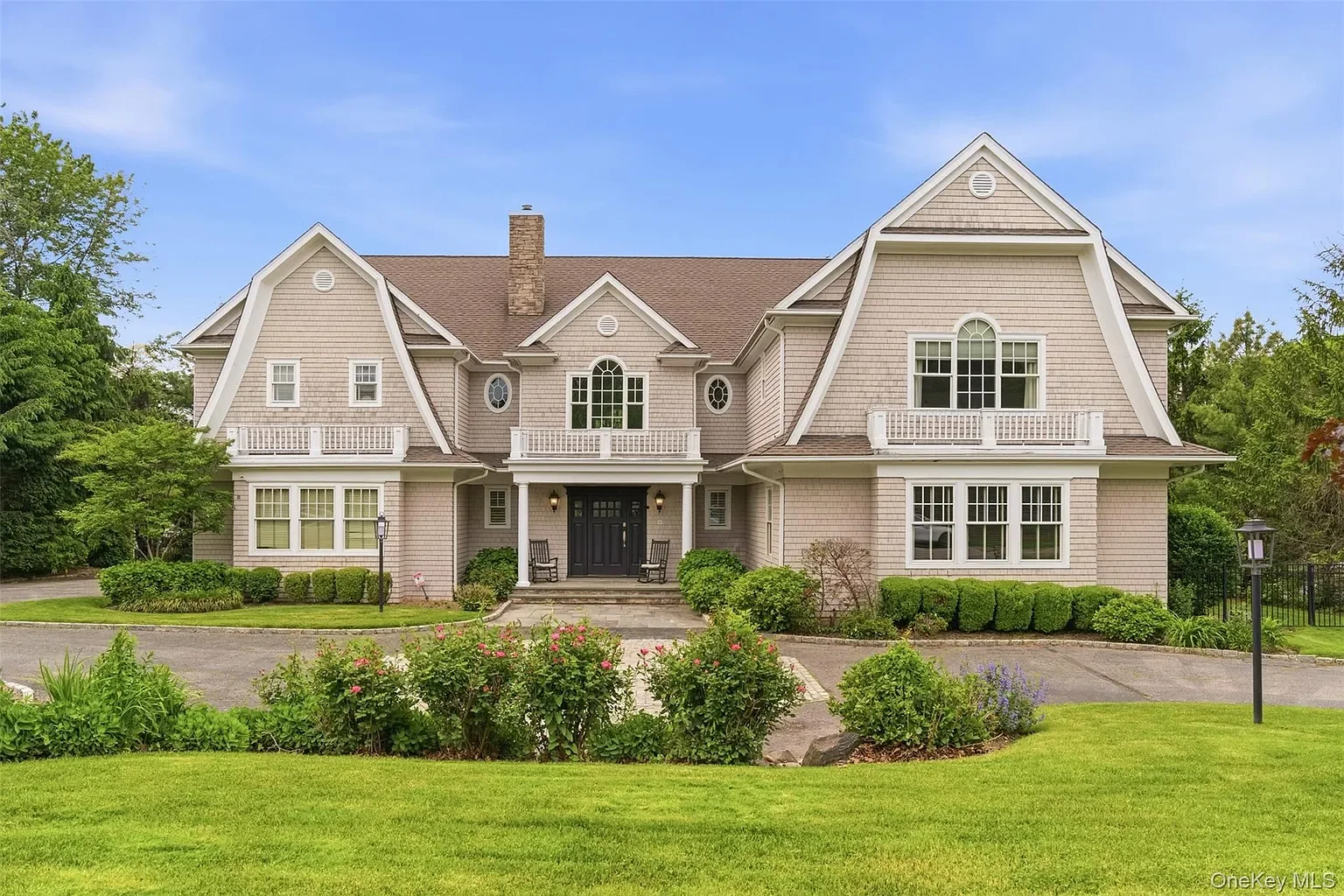 Front view of a large, two-story house with gray siding, multiple windows, and a brown roof, surrounded by greenery and a well-maintained lawn.
